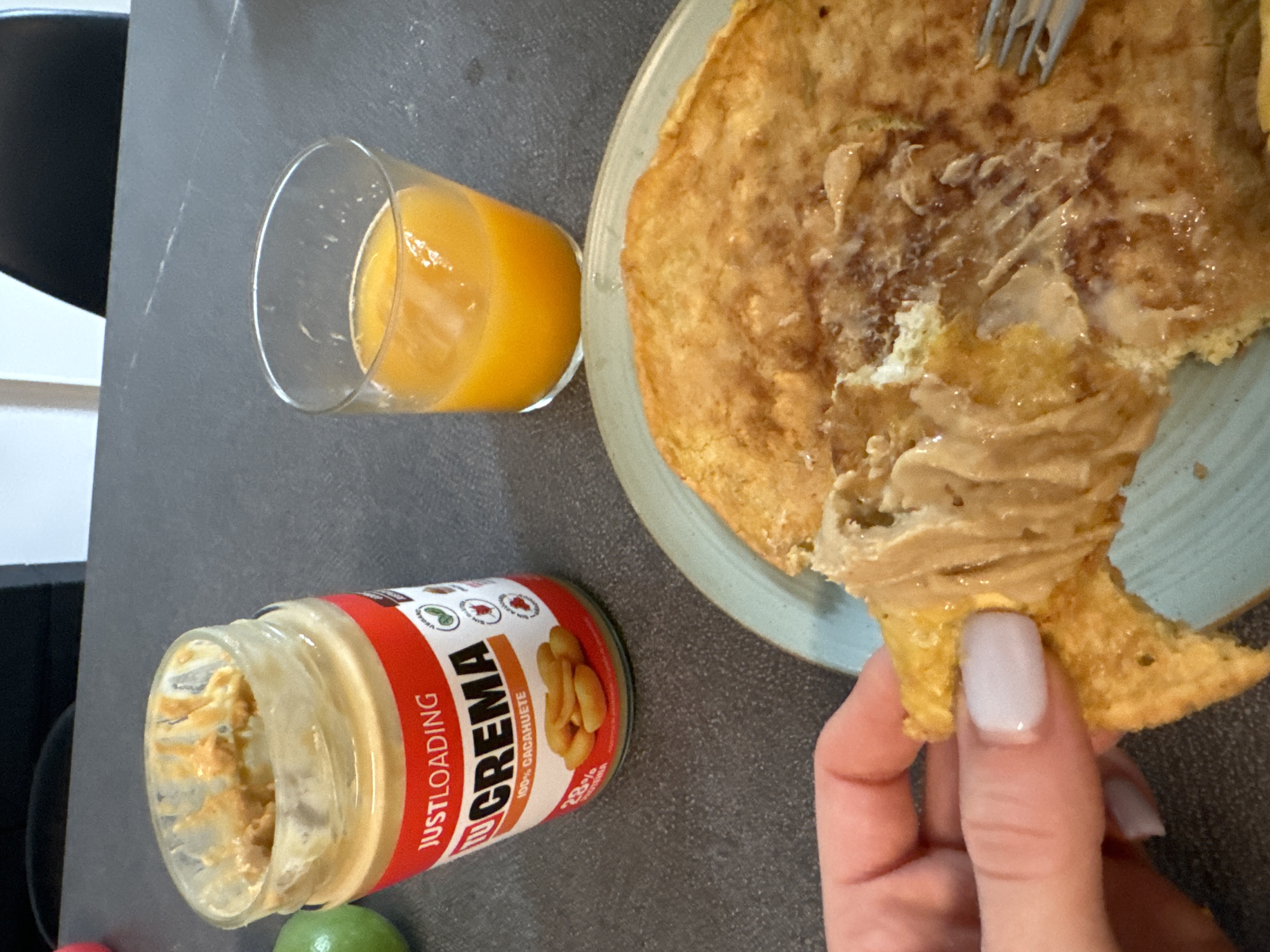 A close-up shot shows a breakfast scene on a grey countertop. In the foreground, a jar of peanut butter labeled "JUSTLOADING LUCREMA 100% CACAHUETE" is open, with some peanut butter remaining inside. To the left, a glass filled with orange juice sits next to the peanut butter jar. In the background, a pancake, partially eaten and spread with peanut butter, rests on a light blue plate. A fork is visible resting on the pancake. A hand with light pink nail polish is holding a piece of the pancake. The lighting suggests it is daytime, possibly indoors.
