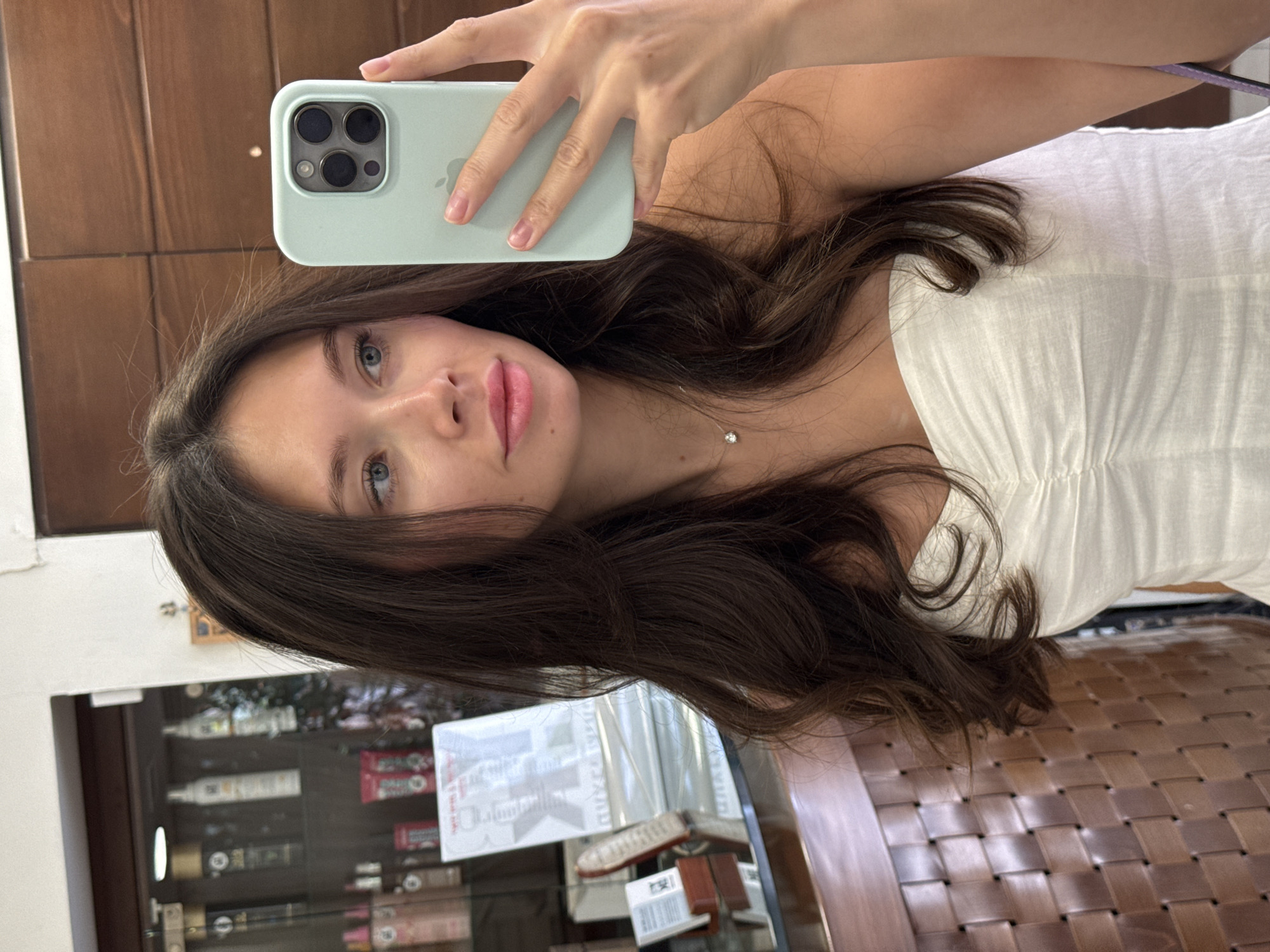 The image captures a mirror selfie of a young woman with long, dark brown, wavy hair and blue eyes, wearing a white strapless or off-the-shoulder top. Her right hand, with natural nails, holds a light green iPhone, its triple camera array and "iPhone" logo clearly visible on the back of the case. A delicate silver-colored necklace with a small, clear gem adorns her neck. She looks directly towards her reflection with a neutral expression and slightly parted, pink-tinted lips.
The background, reflected in the mirror, suggests an indoor commercial setting, likely a salon or beauty store in Petah Tikva, Israel. To the left, a display cabinet filled with various bottles and packages, presumably cosmetics or hair products, is visible. Some items have red and white packaging, and numerous cylindrical bottles are arranged on shelves. Small white labels with unreadable text are attached to the cabinet. Warm brown wooden panels form part of the background, along with a richly woven, dark brown material, possibly part of a chair, bench, or decorative panel. On the far left, a white door frame or trim is partially visible. The lighting is bright and even, characteristic of an interior space.