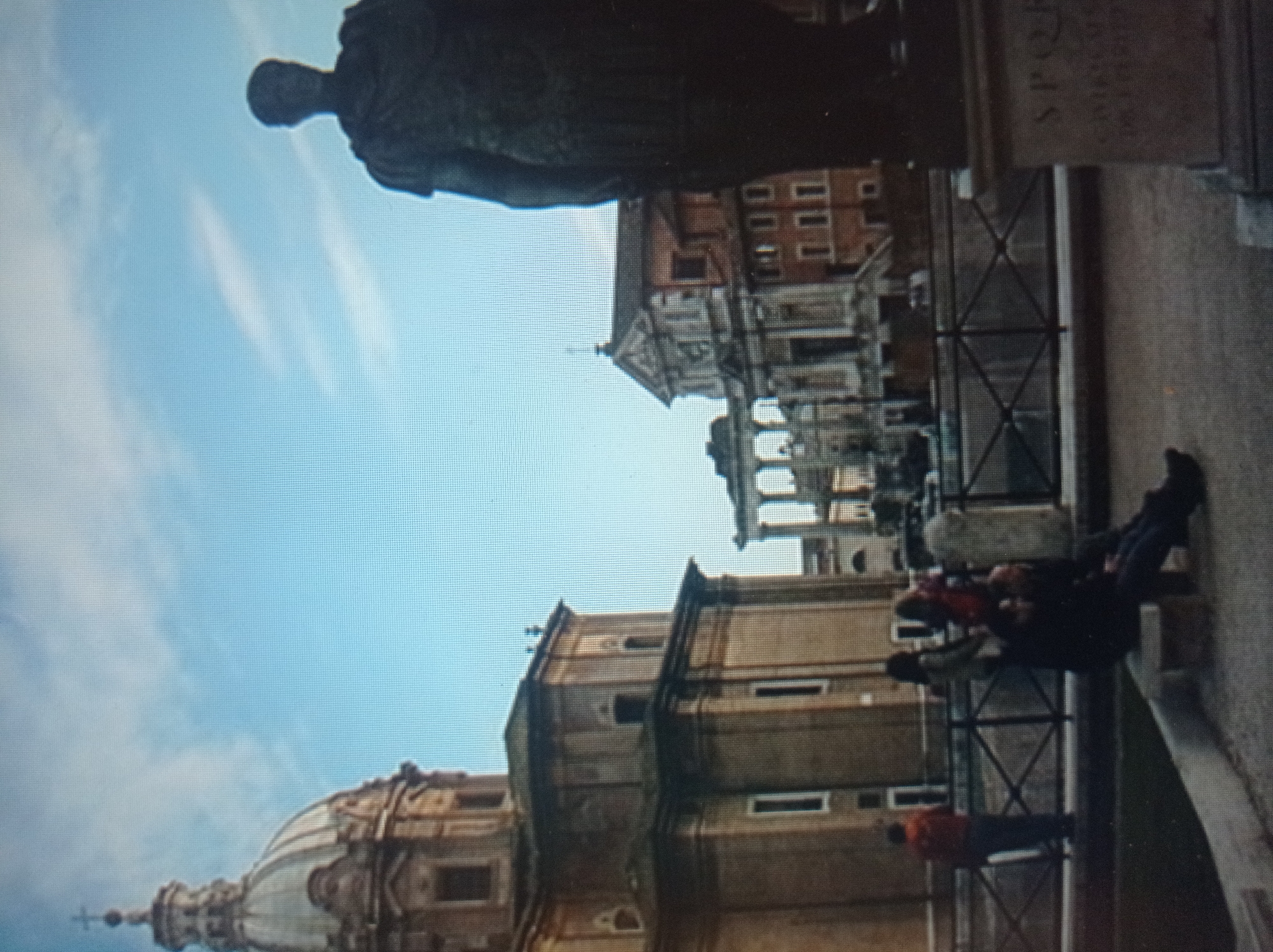 The image depicts a wide shot of an outdoor scene, featuring a grand architectural dome on the left and a large statue of a person in Roman attire on the right. In the middle ground, several people are visible, some standing and some sitting on a ledge. Behind them, more classical architecture is present, suggesting a historical or monumental setting. The sky above is a clear blue with some wispy clouds. The statue on the right has Latin text on its pedestal. The people in the scene are casually dressed and appear to be tourists or visitors. The environment seems to be a public space, possibly a park or square, with a fence in the foreground. The overall atmosphere is one of historical grandeur and leisure. The location of Mukachevo, Ukraine is not evident from the image.
