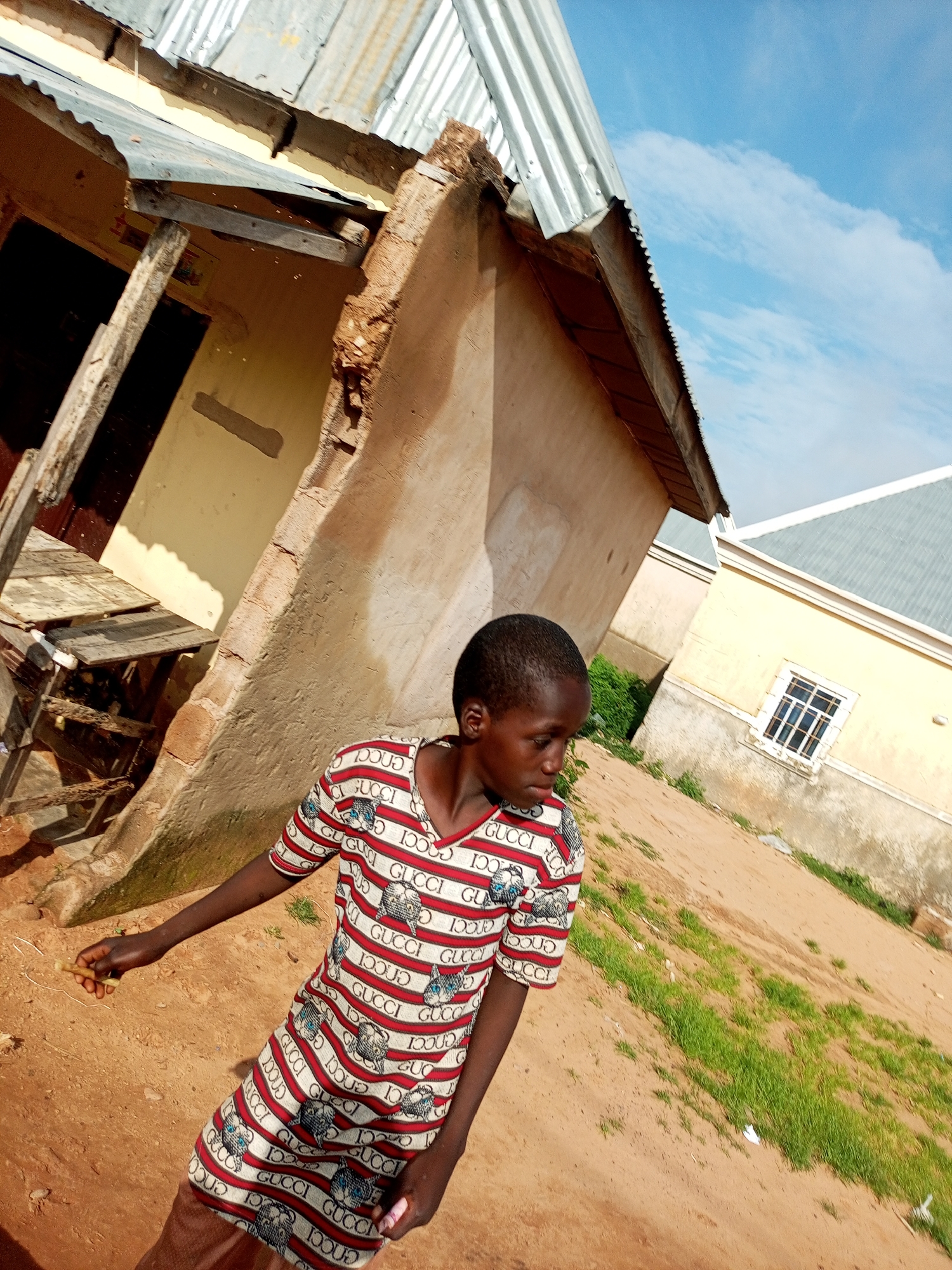 In an outdoor setting, a young person stands in front of a partially constructed or dilapidated building. The building has a corrugated metal roof and pale yellow walls, with exposed wooden beams and some crumbling plaster. To the left, a wooden structure resembling a table or shelf is visible. The ground is covered in dry, sandy soil with patches of sparse green grass. In the background, another building with a similar roof and yellow walls, featuring a white-framed window, is partially visible. The sky above is a clear blue with a few wispy white clouds. The young person is wearing a striped t-shirt with a repeating "GUCCI" logo and cat motifs. Their right hand is extended, holding a small, light-colored object. Their gaze is directed downwards and to the right. The scene appears to be in a rural or semi-urban area, with a focus on the informal construction and natural elements of the environment.