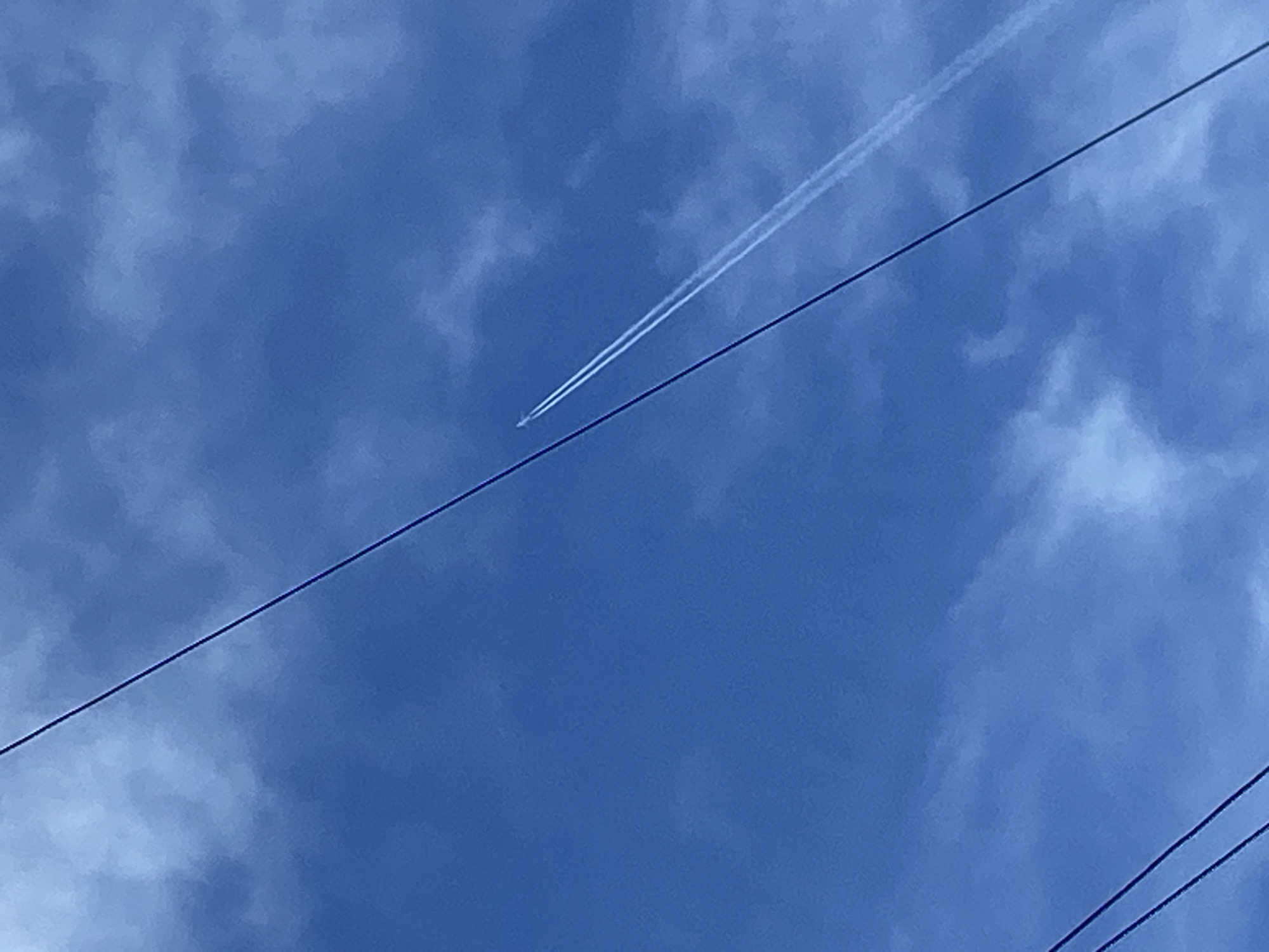 A daytime, low-angle shot of a blue sky with scattered clouds. An airplane is flying, leaving a contrail. Several power lines run diagonally across the frame. The location cannot be ascertained from the image.