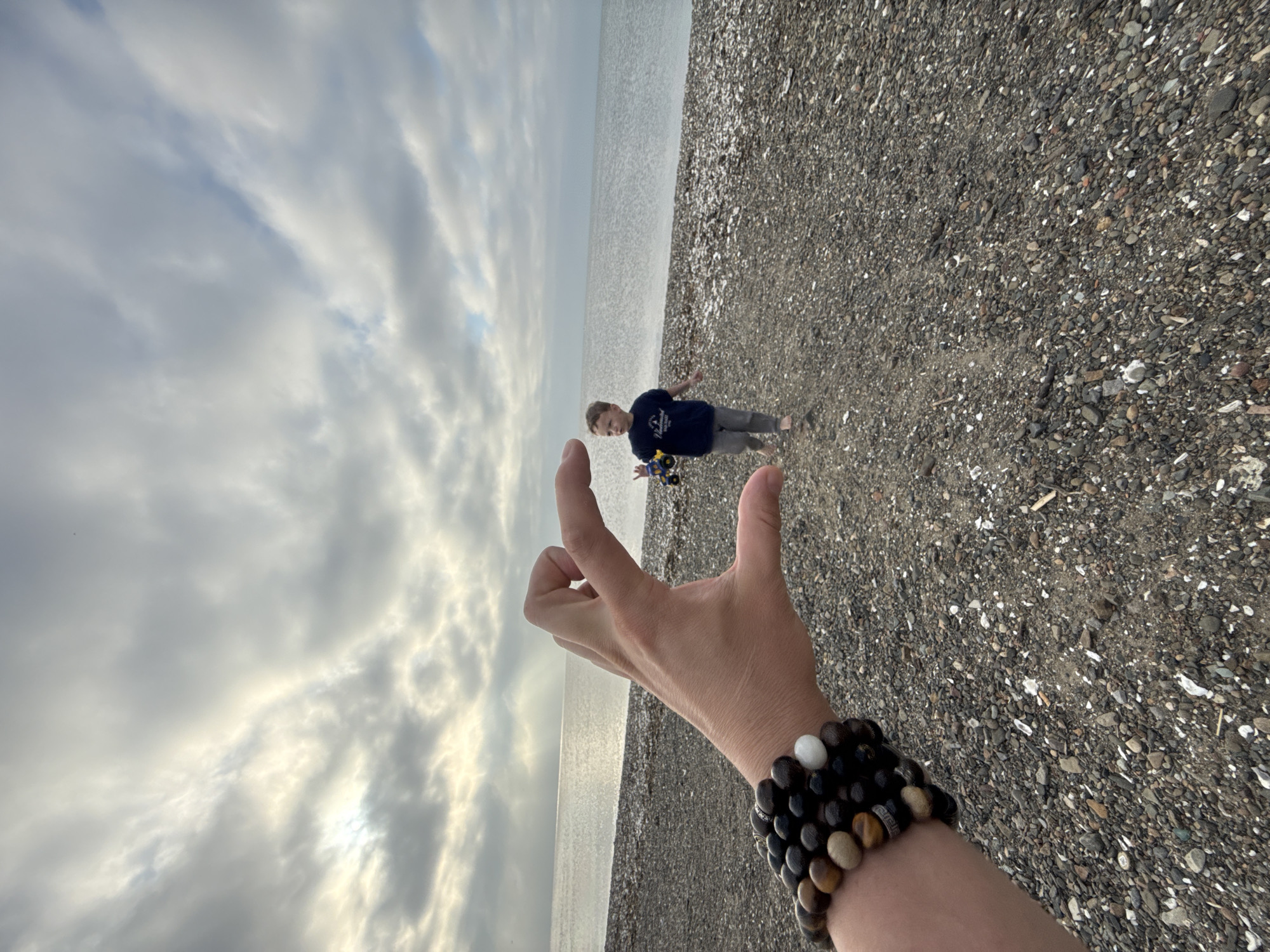 An adult's hand, adorned with bracelets, is in the foreground, forming a "C" shape. Beyond the hand, a young boy walks on a pebble beach toward the ocean. He is wearing a dark blue t-shirt and gray pants and is holding a toy truck. The sky is overcast with dramatic clouds. The scene appears to be a beach in Vladivostok, Russia.