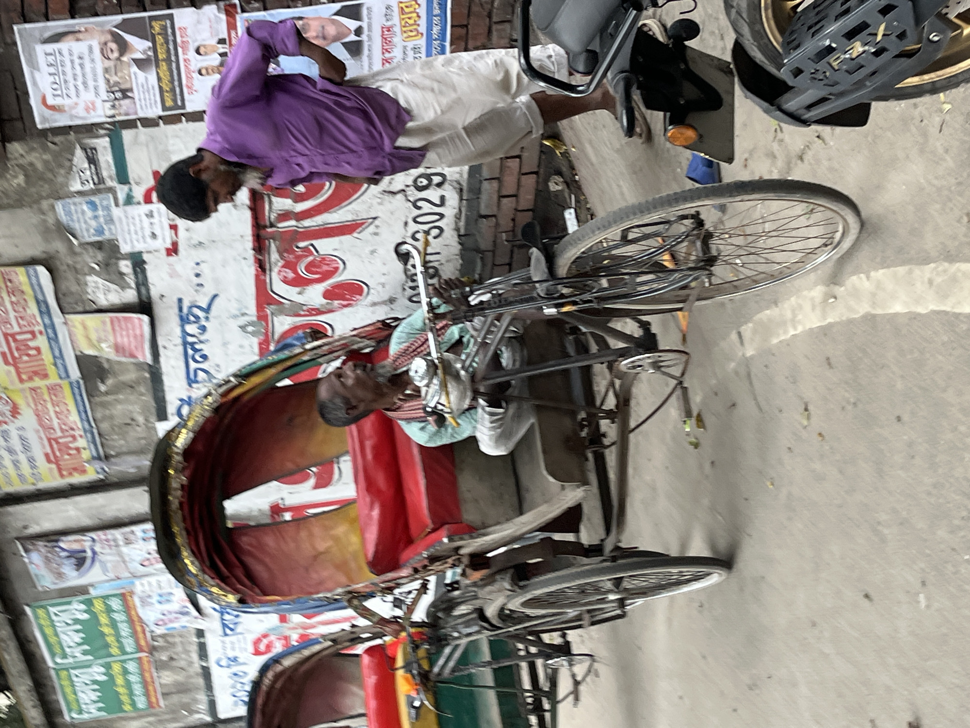 The image shows an urban scene likely in Dhaka, Bangladesh, with a man standing near a rickshaw on a street lined with posters and advertisements. Another man is sitting on the rickshaw. A motorcycle is also visible. The surrounding environment appears to be a bustling urban area, given the number of posters and the presence of both pedestrian and vehicular traffic.