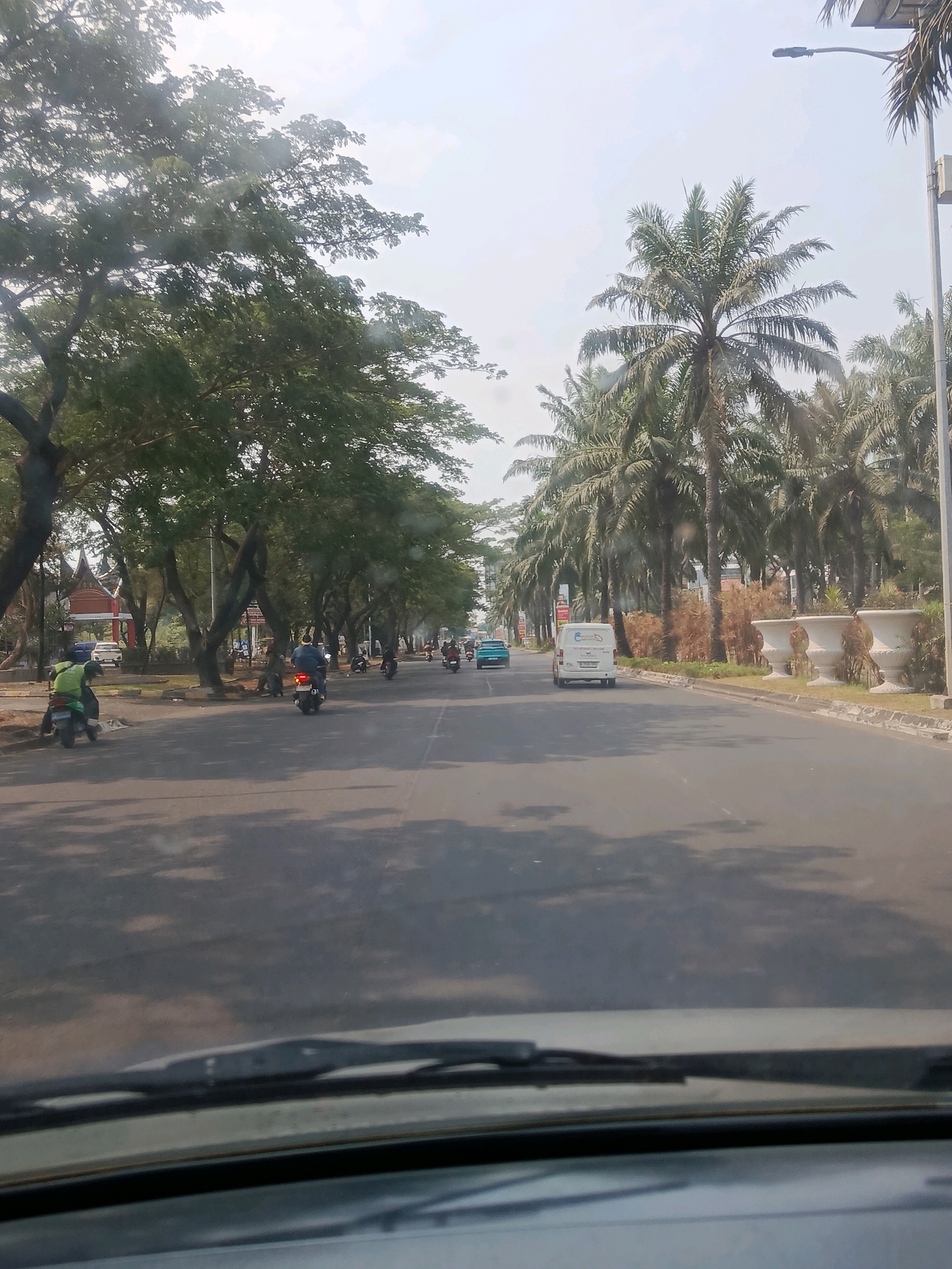 The image depicts a street scene in the city of Bekasi, Indonesia. The perspective is from inside a vehicle, looking forward through the windshield. The road is paved and appears to be a multi-lane street lined with trees on both sides. On the left, there are large trees with dense foliage, casting shadows on the road. Further down this side, a reddish-brown building with intricate roofing and an archway is visible. Several motorcycles are on the road and on the side, with some riders visible. A person in a green jacket is seated on a motorcycle on the left.
On the right side of the road, a row of tall palm trees stands out against a bright, hazy sky. Several large, ornate white planters are placed along the sidewalk. A white delivery van with branding is driving in the right lane. The sky is overcast and bright, suggesting it is daytime. The overall impression is that of a well-maintained urban thoroughfare with significant greenery.