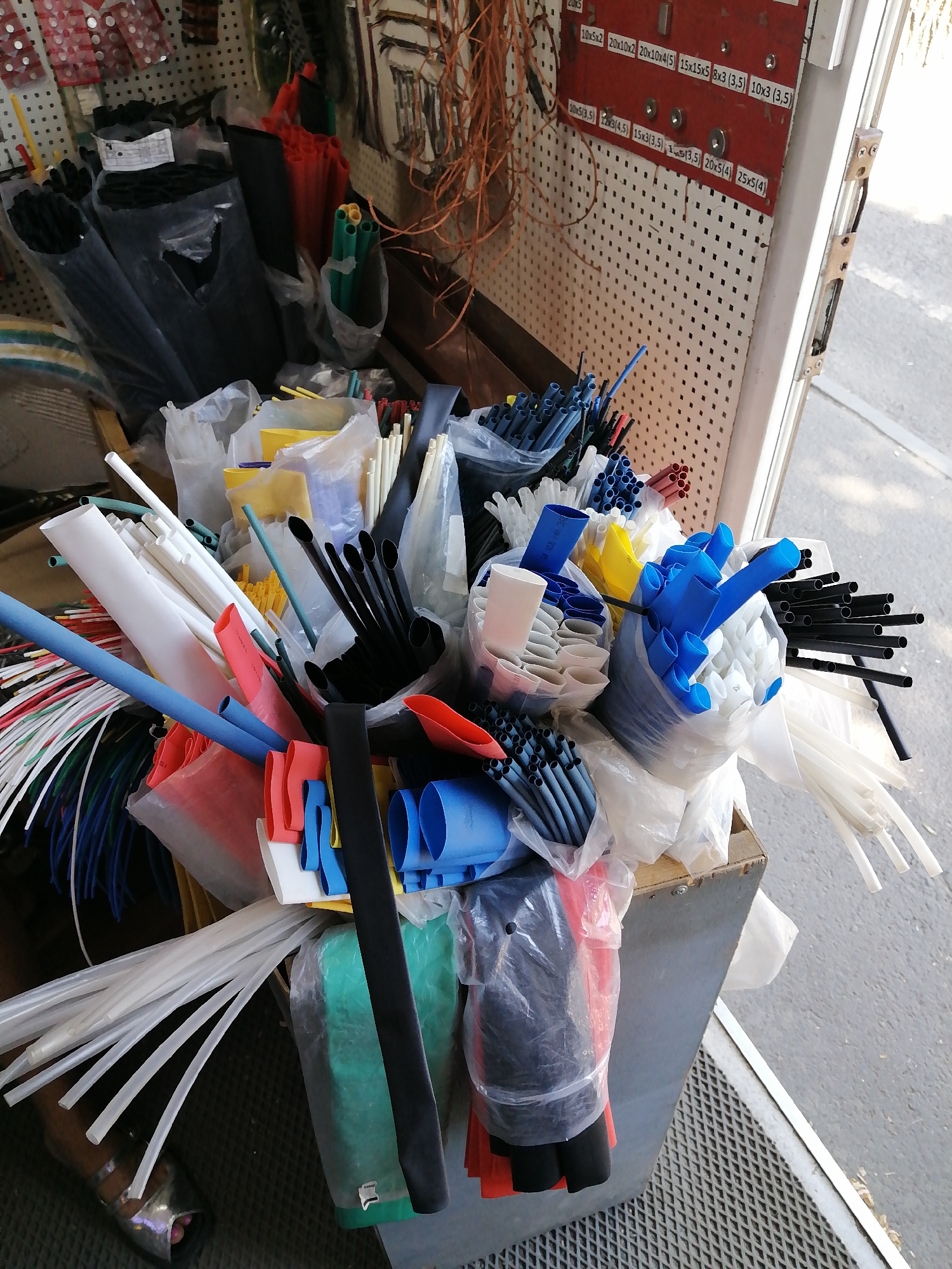 The image depicts an outdoor market stall or workshop area in Rostov-on-Don, Russia. The scene is dominated by a large collection of electrical insulation materials, primarily heat-shrink tubing and wiring protection. These are organized in a somewhat haphazard manner within a metal bin and on shelves in the background. The materials are in various colors, including black, white, blue, red, yellow, and green. Some of the tubing is neatly bundled, while other pieces are loosely packed or spilling out of their containers. In the background, a pegboard displays various electrical components and tools, with labels indicating sizes and specifications, suggesting a professional or hobbyist setting. The overall impression is one of a busy, functional workspace where a variety of electrical supplies are stored and displayed for sale or use.