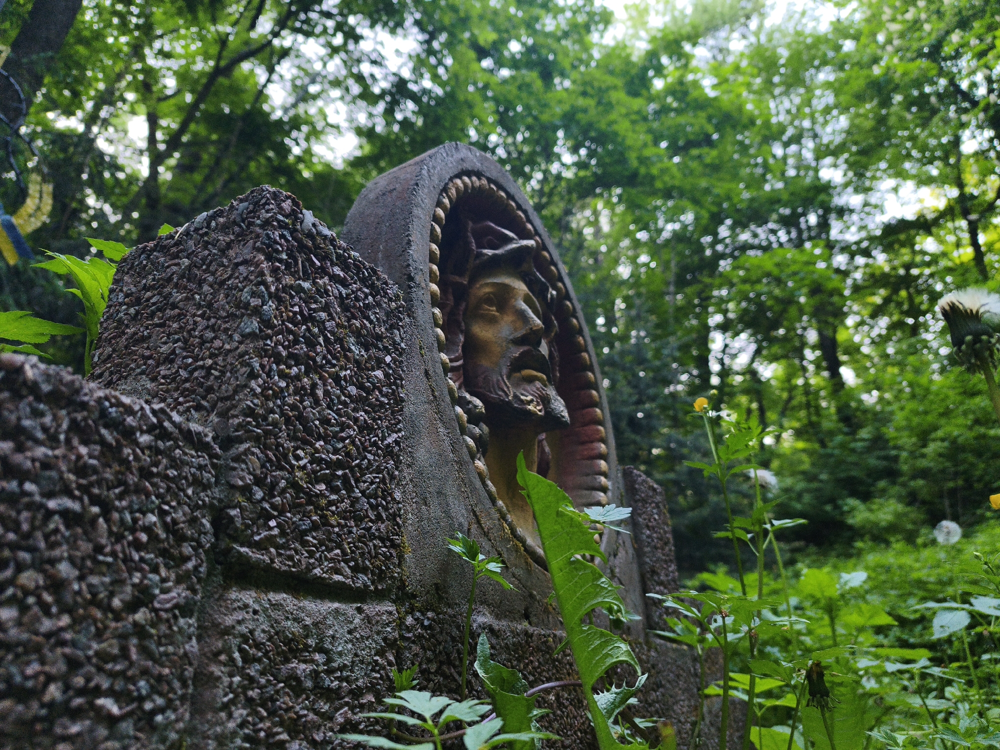 A close-up shot captures a stone statue of a man's head within a rounded frame, likely of religious significance, possibly Jesus Christ, given the head covering and beard. The statue appears weathered and is set within a structure made of rough, pebble-like material. Lush greenery surrounds the statue, suggesting it's located in a garden or wooded area. The background reveals a dense forest, filled with tall trees and various plants. The scene evokes a sense of peace and solitude. The statue is located in Kopychyntsi, Ukraine.