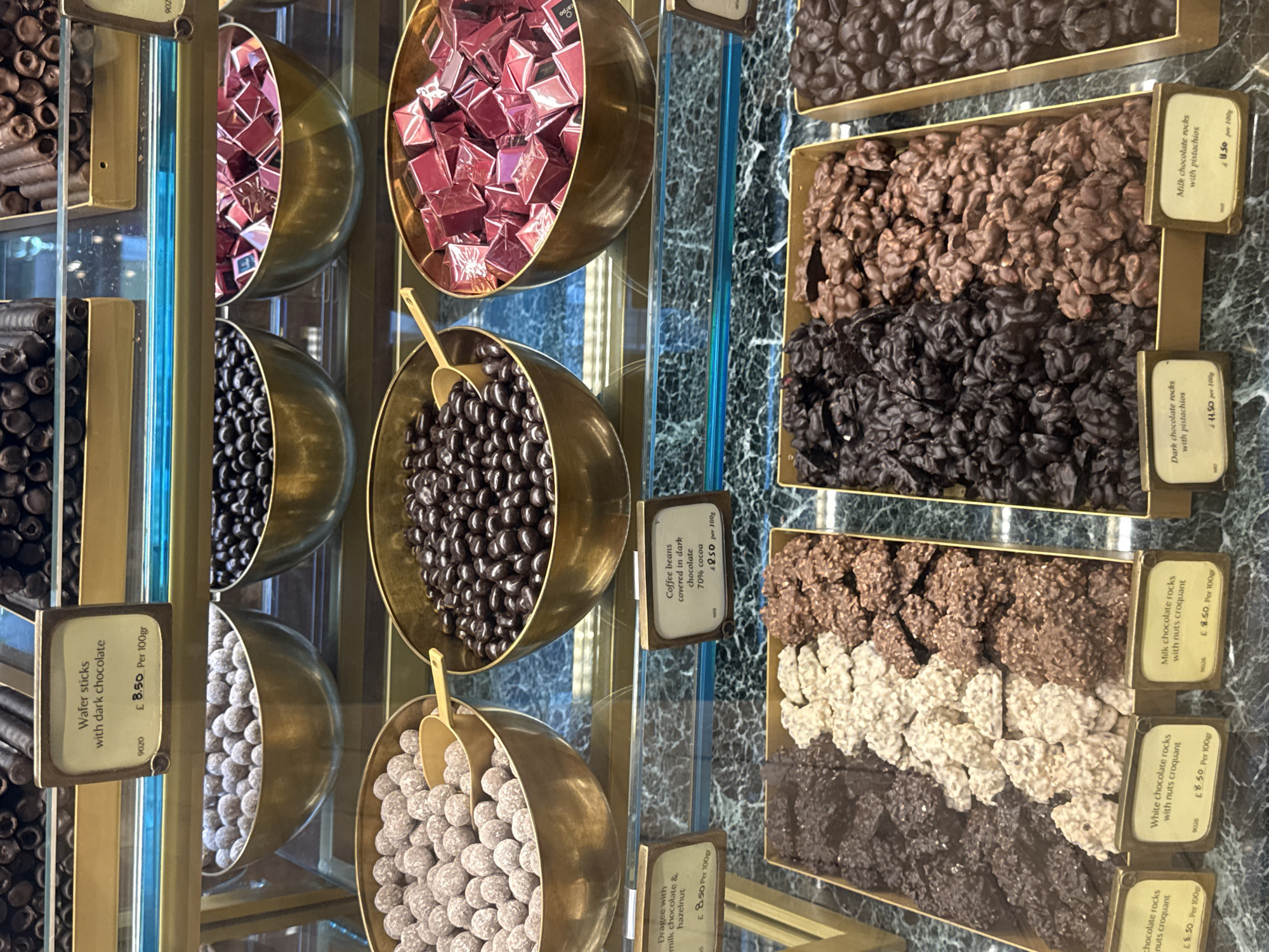 This media file presents a vibrant, close-up view of a luxury chocolate display, characteristic of a high-end confectionery shop or a patisserie in London. No people are visible, emphasizing the carefully arranged products.
The scene showcases an array of chocolates and confections, meticulously displayed in ornate, brass-colored bowls and elegant gold-rimmed trays. On the left, several bowls are vertically arranged, holding dark chocolate wafer sticks, milk chocolate and hazelnut dragees, and coffee beans covered in 70% cocoa dark chocolate. Small golden scoops rest in some of these bowls.
To the right, flat trays present various types of chocolate rocks, including milk chocolate rocks with pistachios, dark chocolate rocks with pistachios, and different varieties of milk, white, and dark chocolate rocks with nuts croquant. The chocolates are neatly separated into distinct sections, highlighting their textures and ingredients.
Each product features a small, framed price tag providing its name, weight (per 100g/gr), and price in British Pounds (£). Visible text includes: "Wafer sticks with dark chocolate £ 8.50 Per 100gr", "Dragee with milk chocolate & hazelnut £ 8.50 Per 100gr", "Coffee beans covered in dark chocolate 70% cocoa £8.50 per 100g", "Milk chocolate rocks with pistachios £ 11.50 per 100g", "Dark chocolate rocks with pistachios £ 11.50 per 100g", "Milk chocolate rocks with nuts croquant £ 8.50 Per 100gr", "White chocolate rocks with nuts croquant £ 8.50 Per 100gr", and "chocolate rocks nuts croquant £6.50 Per 100gr". The display rests on a dark, marbled surface, with reflective glass and gold-colored metal elements enhancing the luxurious setting. The meticulous arrangement and premium pricing suggest a high-quality establishment.