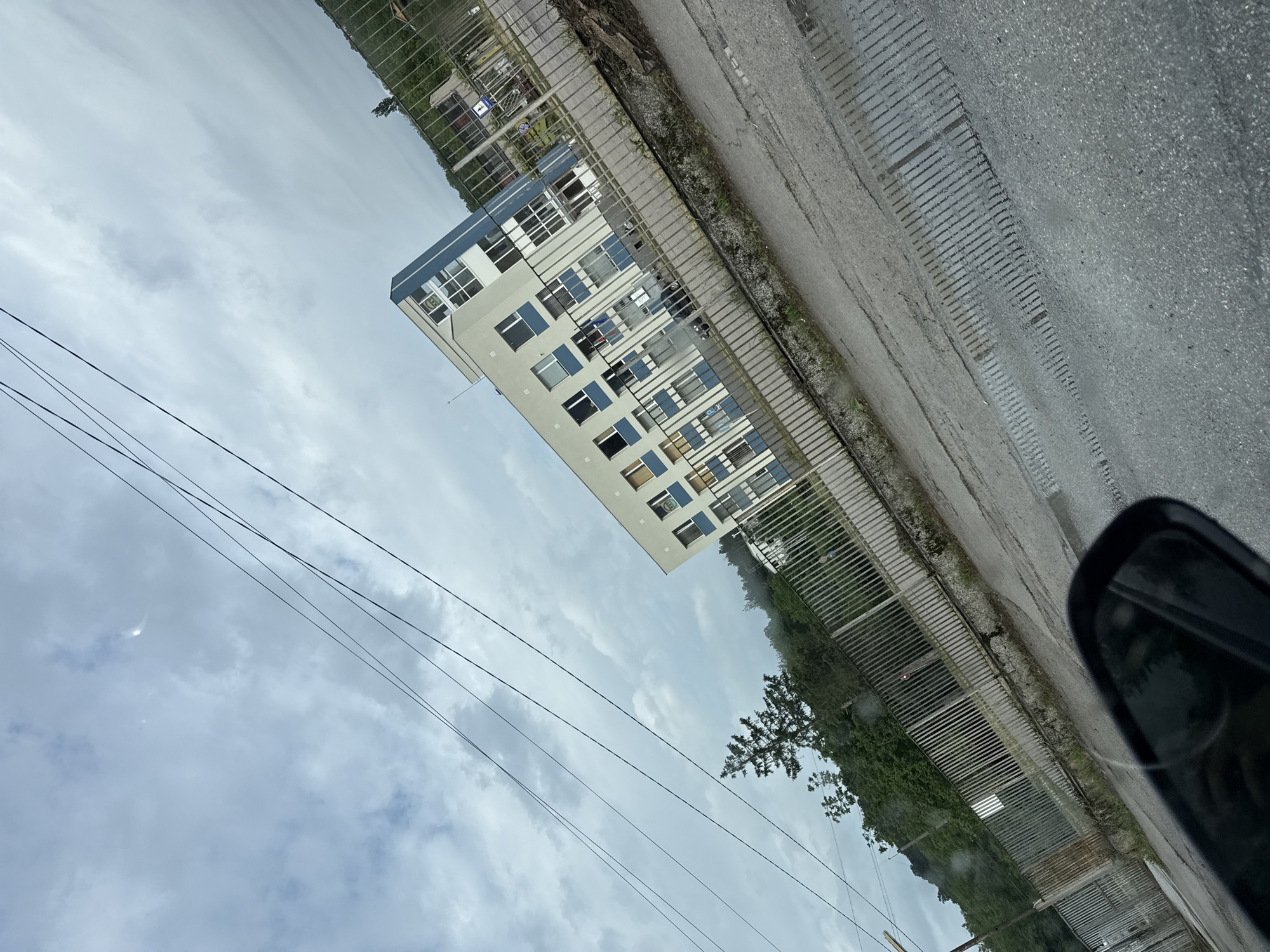 A low-angle shot shows a modern building with white walls and blue window accents, likely residential or commercial, standing near a sidewalk and road in Unknown, Unknown. The sky is overcast with gray clouds, and power lines stretch across the upper-left quadrant. A metal fence runs along the perimeter of the building, adjacent to green foliage. A side mirror of a vehicle is visible in the lower right corner, implying the shot was taken from inside a car. The road has visible tire tracks and wet patches, suggesting recent rainfall.