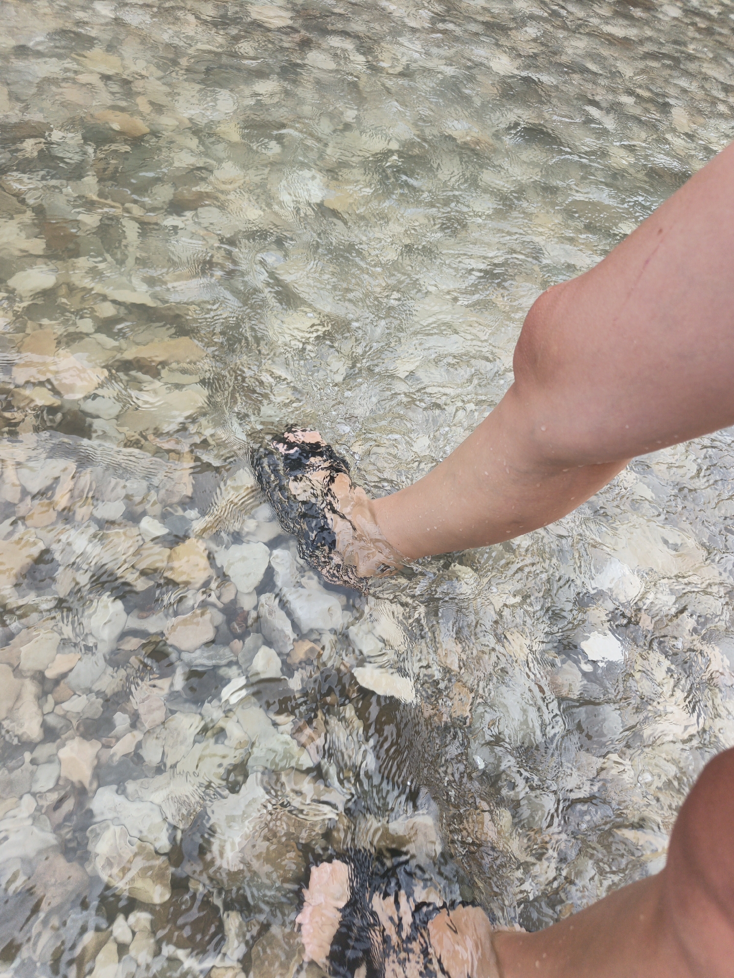 The media file shows a close-up view of a person's legs and feet submerged in shallow, clear water with a rocky bottom. The water is rippling, creating distortions in the view of the pebbles and stones beneath. The pebbles are a mix of light and dark colors, mostly gray, beige, and brown, with some appearing smooth and rounded, while others are rougher.
The person is wearing dark, possibly water-resistant shoes, visible on both feet. The legs are lightly tanned, and small water droplets are visible on the skin of the right leg, indicating recent immersion or splashing. The overall scene suggests an outdoor setting, likely a riverbed or shallow stream in a natural environment. The light appears to be natural daylight, casting subtle highlights on the water's surface and illuminating the pebbles below. There are no visible signs of text or specific landmarks to pinpoint the exact location beyond the general context of a rocky watercourse. The activity appears to be a casual one, possibly wading, cooling off, or exploring a natural water feature. The perspective is from above, looking down at the legs and feet.
