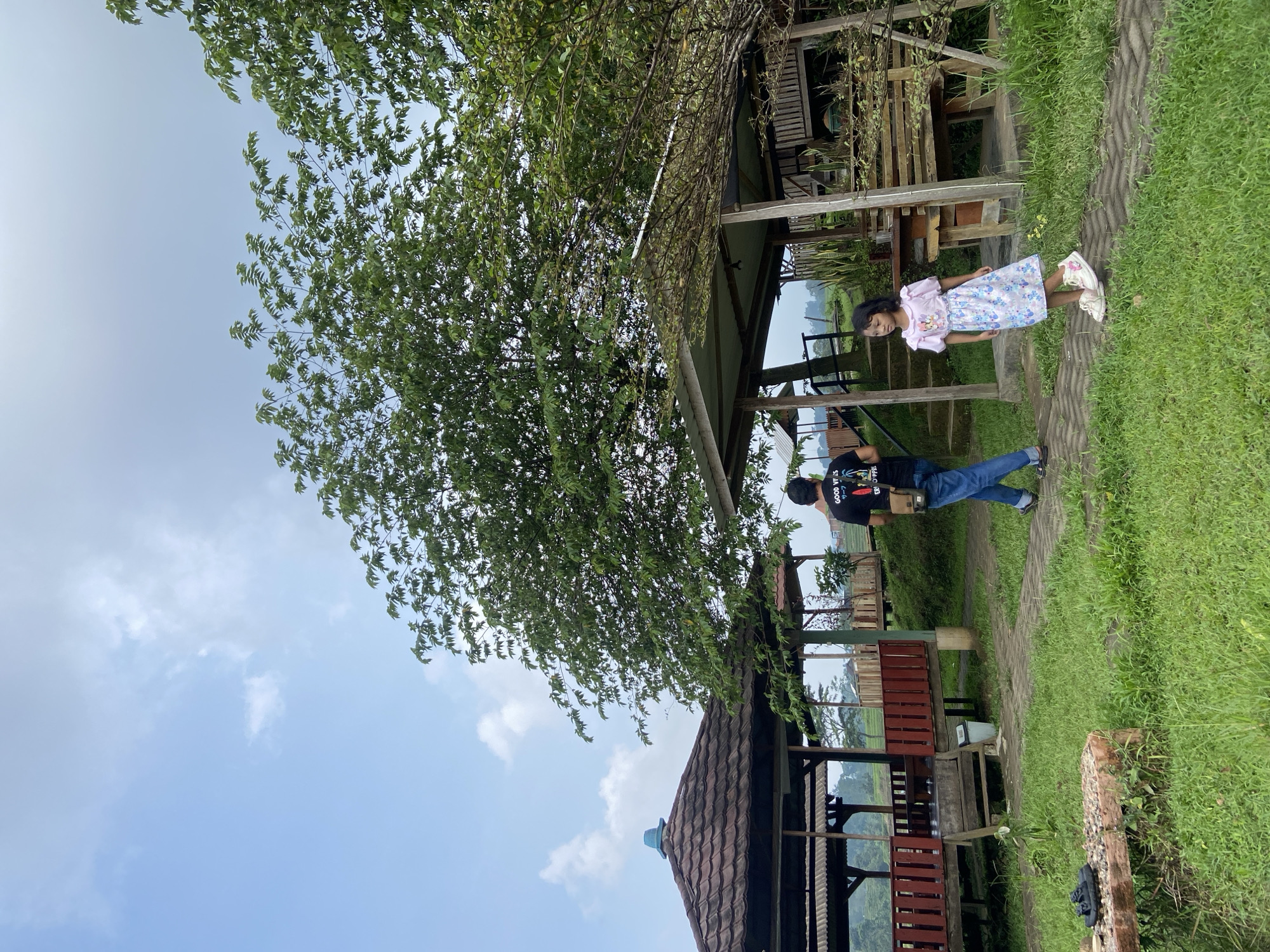 A young girl stands near a man outside of a rustic, partially enclosed wooden structure with a tile roof. The girl wears a light-colored dress and white shoes. The man wears a dark t-shirt, blue jeans, and casual shoes. They appear to be located in a grassy area near a path. The setting appears to be Penanggungan, Indonesia. A large tree provides shade in the foreground, and additional similar structures are visible in the background. The sky is visible above.
