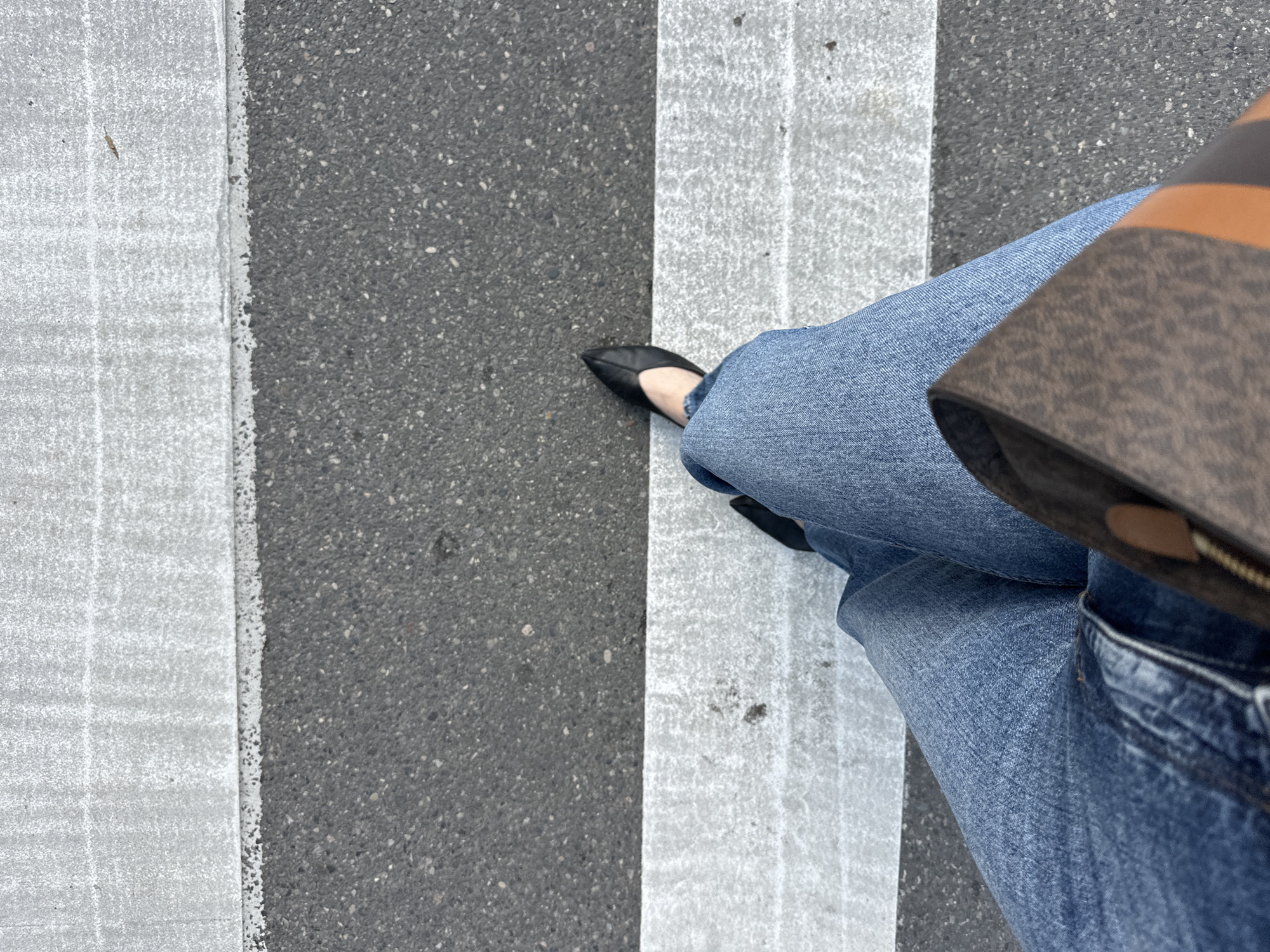 A person is standing on a crosswalk in Batumi, Georgia. The crosswalk is marked with alternating black asphalt and white painted stripes. The person is wearing blue jeans and black flats. A brown purse is partially visible on the right side of the frame. The person is standing on a white stripe. The asphalt shows wear.