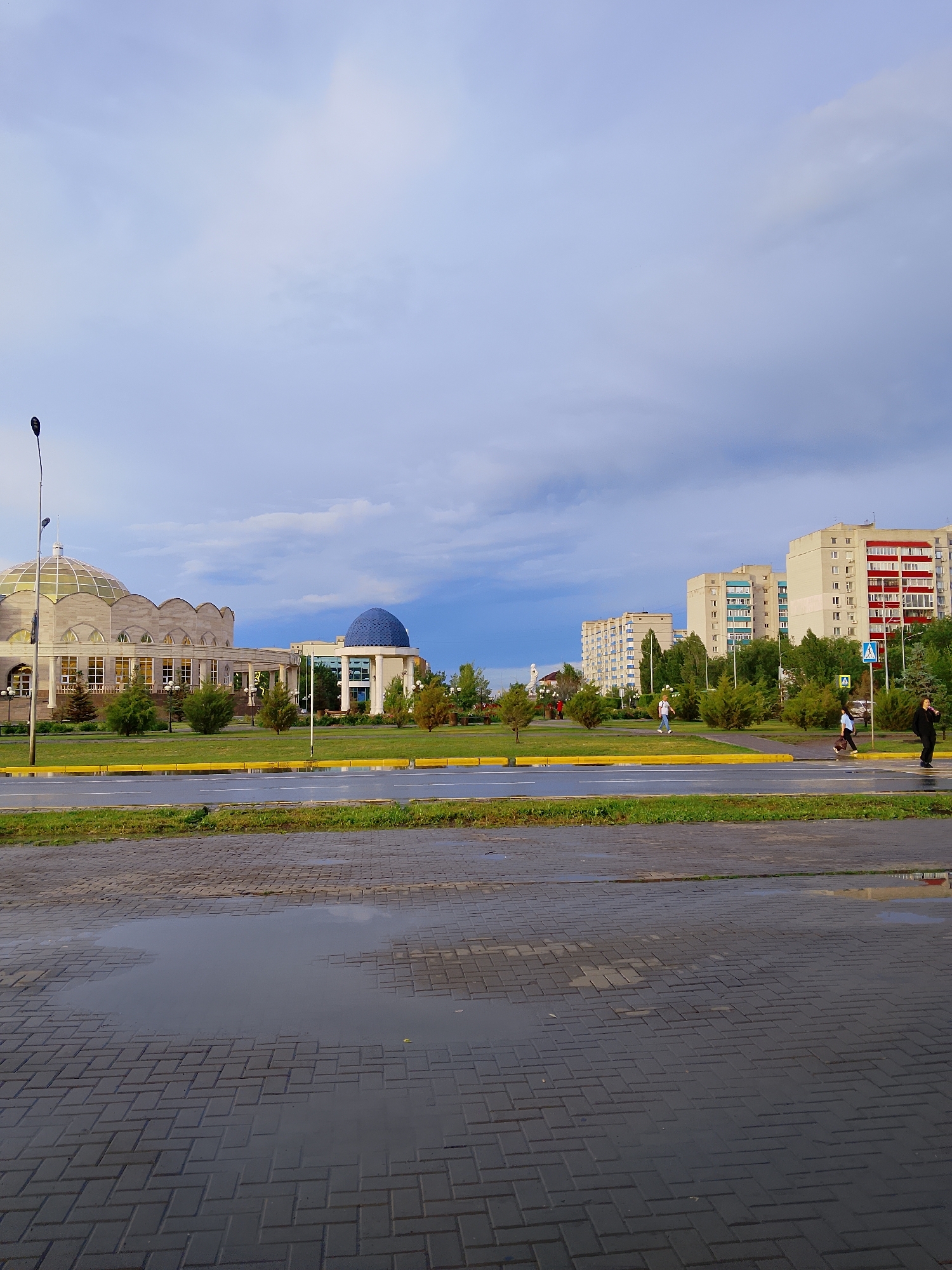 This image depicts an urban scene in Oral, Kazakhstan, following a rain shower. The sky is overcast with dramatic clouds, suggesting either early morning or late afternoon. The wet pavement in the foreground reflects the sky and surrounding environment, indicating recent rain.
The scene features several buildings of varying architectural styles. To the left, a large, curved building with a glass dome and ornate detailing is visible, possibly a cultural center or a government building. Adjacent to it, on the right, is another building topped with a bright blue dome, standing out against the muted sky. In the background, a row of tall, modern apartment buildings with balconies and red and white accents dominates the skyline. Green spaces with trees and manicured lawns are interspersed between the buildings, suggesting a planned urban development.
Several people are present in the image, walking across the street or on the sidewalks. Their presence suggests everyday activity, unaffected by the recent weather. A street lamp stands tall on the left side of the frame, a common urban fixture. There are no visible signs with text. The overall mood is calm and somewhat subdued, likely due to the weather conditions. The reflections in the puddles add a sense of depth and atmosphere to the image.