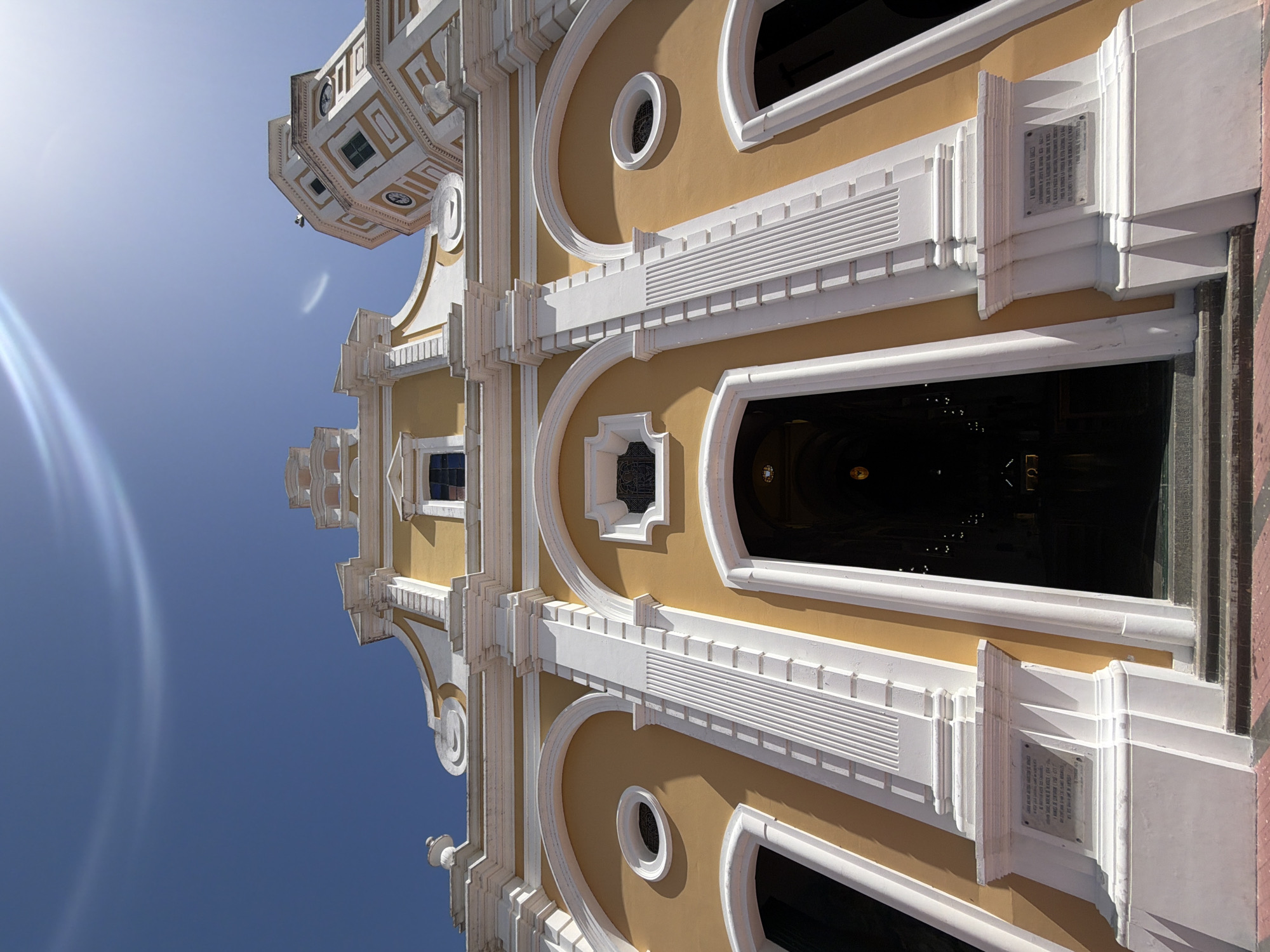 The image shows the exterior of the Ciudad Bolivar Cathedral in Venezuela, taken from a low angle looking up. The architecture is primarily yellow and white with ornate details. The cathedral entrance is visible, leading into a dark interior. A tower structure is seen at the top left of the frame. The sky is clear blue.