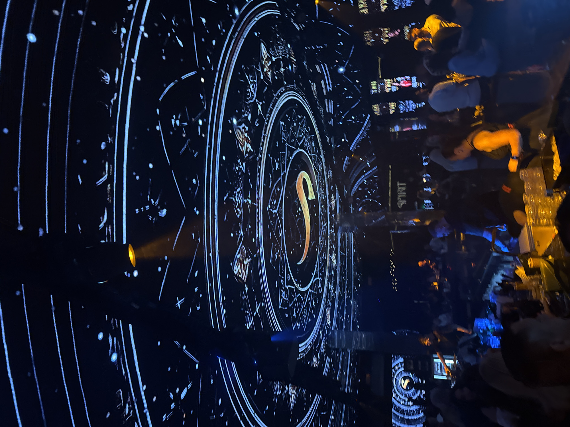 The image depicts a crowded indoor bar or club setting in Plovdiv, Bulgaria. The focus is on a large, circular projection or display behind the bar, featuring a cosmic design with astrological elements, including concentric circles, constellations, and a prominent "S" logo illuminated in warm light. The crowd consists of numerous people, some interacting at the bar and others standing or moving around. A variety of drinks are visible, suggesting a vibrant nightlife scene. The lighting is dim with blue and yellow tones, typical of a nightclub atmosphere. The "SPIRIT" brand name is visible on one of the surfaces in the background.