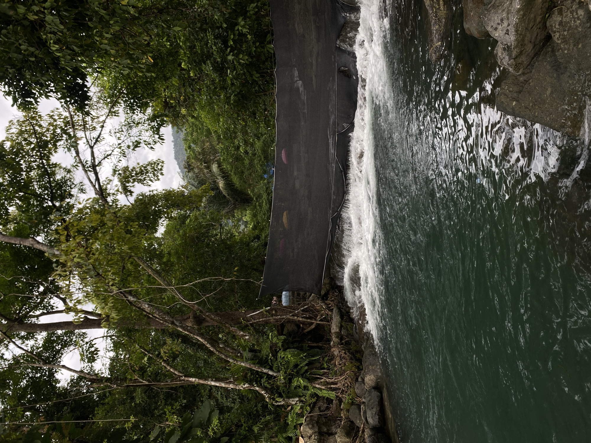 The image presents a scenic view of a small waterfall and surrounding foliage in what appears to be a natural or park-like environment. The waterfall is the prominent feature, with water cascading over rocks into a pool below. The water appears clear and reflects light, creating a shimmering effect. The waterfall is partially obstructed by a dark screen or netting. Lush vegetation, including various trees, ferns, and shrubs, frames the waterfall. The sky is overcast, suggesting a cloudy day. The image does not depict any people or specific actions. The location of the scene is in Pudeng, Indonesia.