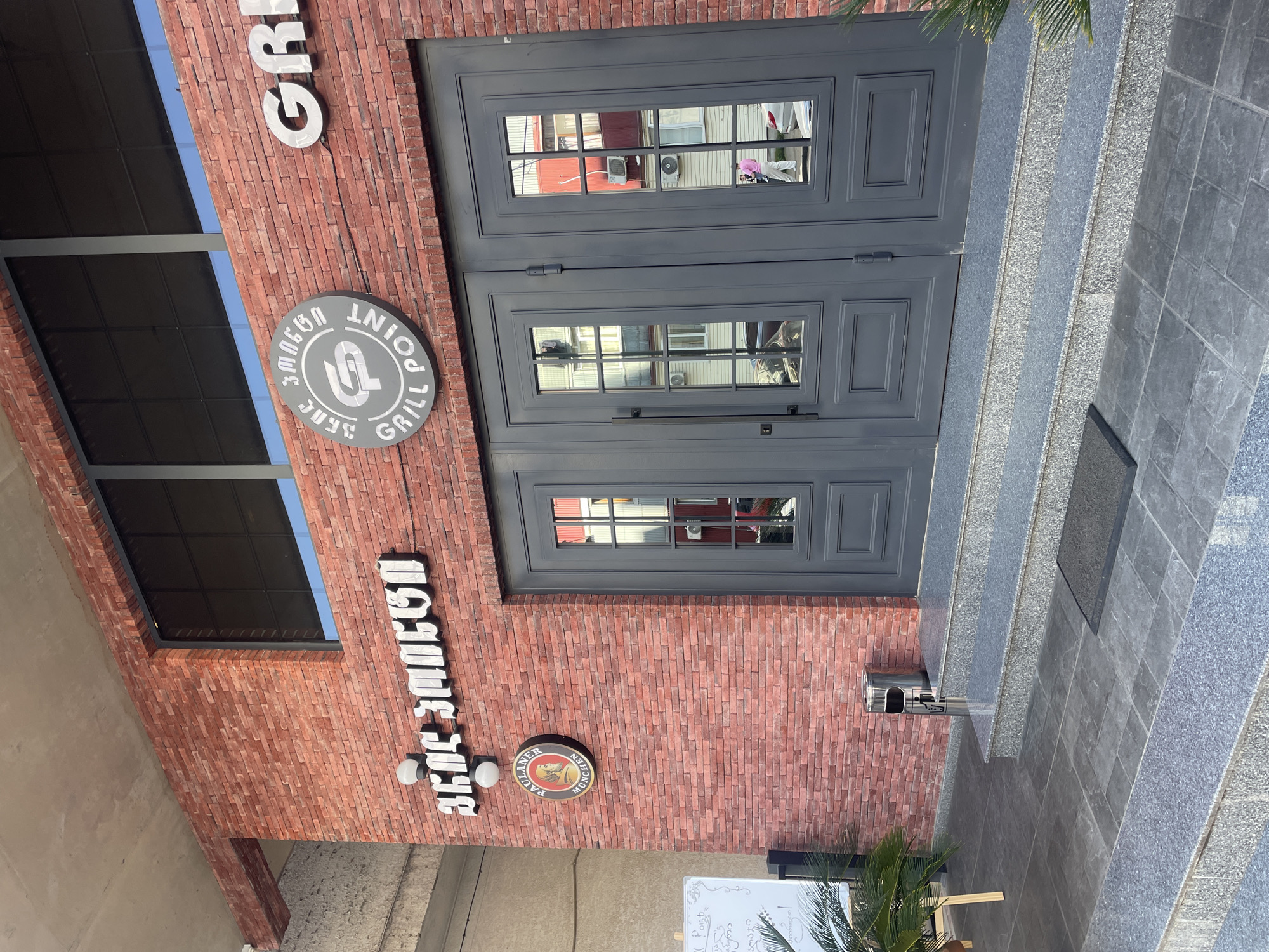 The image shows the entrance to "Union Point Grill" in Batumi, Georgia. The building has a red brick facade. Two signs are visible: one circular with the restaurant's name in Georgian and English, and another vertical sign spelling out "Beer Garden." The entrance features a set of gray double doors with glass panels. A set of gray granite stairs leads up to the doors. An ashtray is mounted on the wall next to the stairs. A whiteboard and palm tree are in the foreground. The image is taken from a slightly low angle.