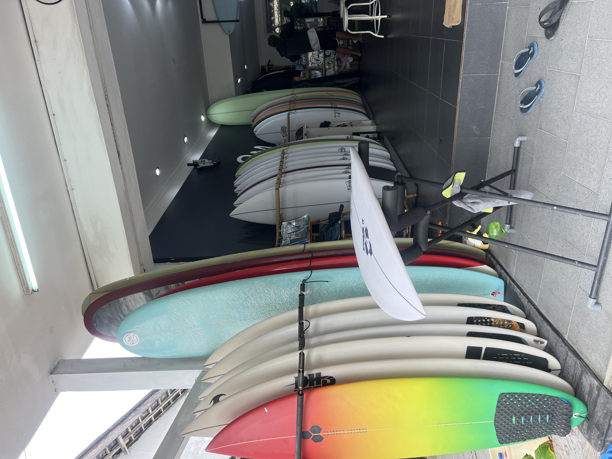 This image depicts a surf shop in Kuta, Indonesia, showcasing a variety of surfboards displayed both inside and outside the establishment. The foreground features a vibrant gradient surfboard with red, yellow, and green hues, standing upright and attached to a black rack. Several other surfboards, predominantly white with subtle colored stripes or designs, are neatly arranged in a rack behind it. One light blue surfboard with a textured surface is prominently visible, leaning against a wall. Further inside the shop, more surfboards, including a tall, light green one, are displayed. The shop has a black wall with some minimalist graphics and a counter area. A person wearing shorts and a jacket is visible in the background, standing near a shelf stocked with items. The floor is tiled with dark gray square tiles. Outside the shop entrance, blue flip-flops and a black pair of flip-flops are scattered on the tiled pavement. The environment appears to be an open-air storefront.