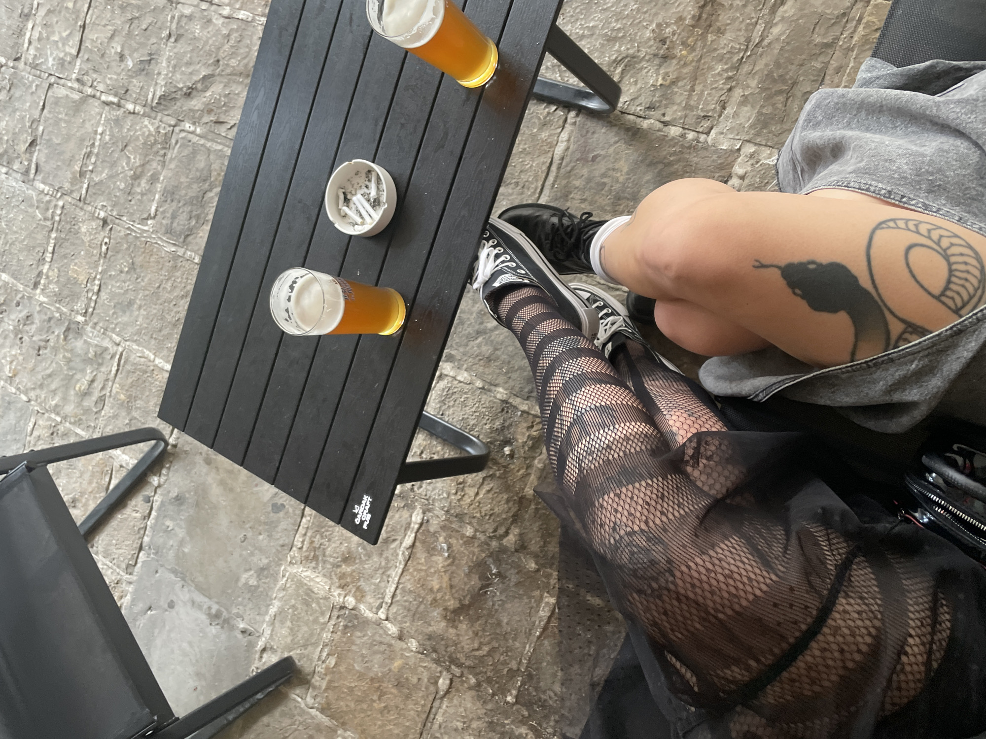 A high-angle shot shows a black table with two glasses of beer and a full ashtray on a stone-paved ground. Two people are seated at the table. One person has a snake tattoo on their thigh and is wearing a grey top, white socks, and black shoes. The other person is wearing fishnet stockings, black sneakers, and a black garment. The table is surrounded by chairs and has a "OUTDOOR CRAFT" logo visible on one corner. The location is the city of Budva, Montenegro.