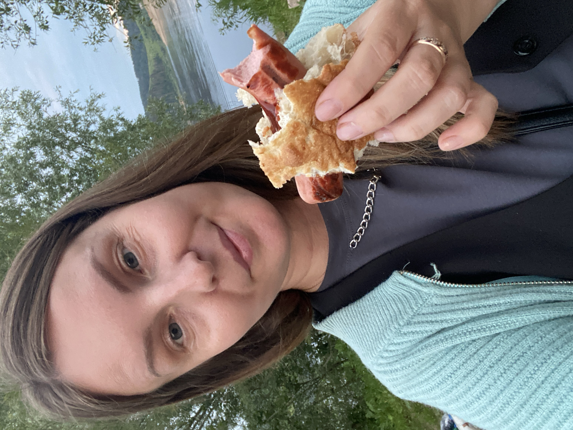 A woman with long brown hair and blue eyes is holding a partially eaten hot dog bun with a sausage in it. She is wearing a light blue zip-up sweater over a dark gray t-shirt and a silver chain necklace. The woman is smiling and looking at the camera. In the background, there is a body of water, trees, and hills. The location is the city of Noviy, Russia.