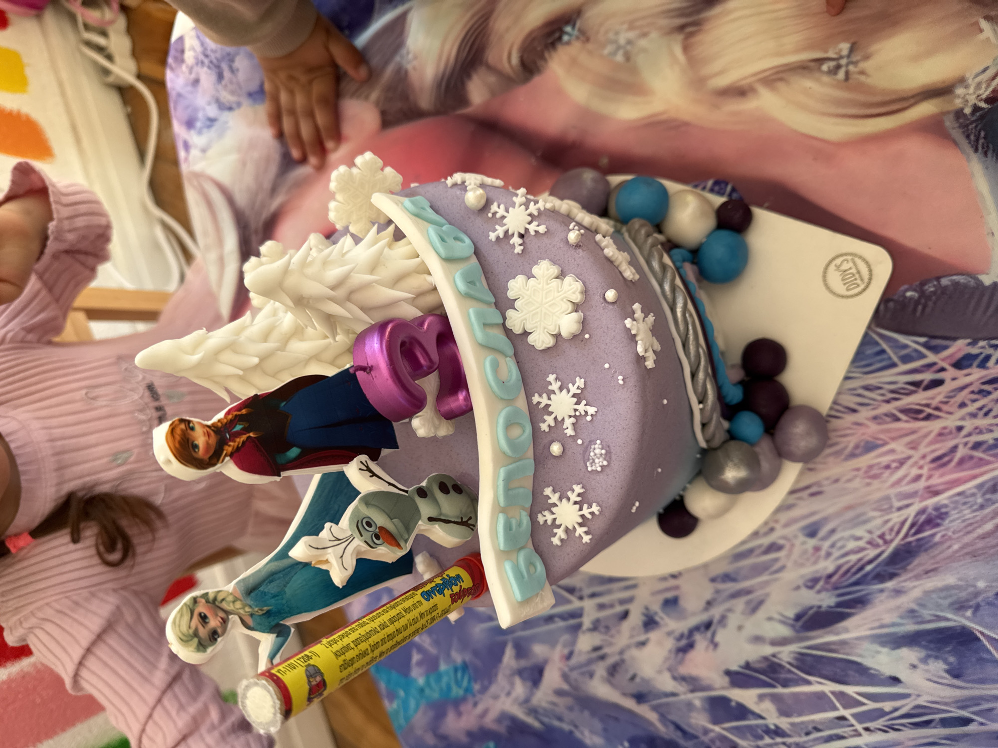 A birthday cake with a Frozen theme is the focal point of the image. The cake is purple and decorated with white snowflakes and blue lettering spelling out "Beроника" (Veronika), and white snow-covered trees. Character images from the movie "Frozen" are on sticks inserted into the cake. A child's hand rests on a backdrop that mimics the "Frozen" landscape behind the cake, and a child's arm and head are visible on the left side of the image. A yellow roll of candy is sticking into the cake. The cake sits on a white cake board. The words "DIDY'S" can be seen on the board. I am unable to determine if the image was taken in Sofia, Bulgaria.