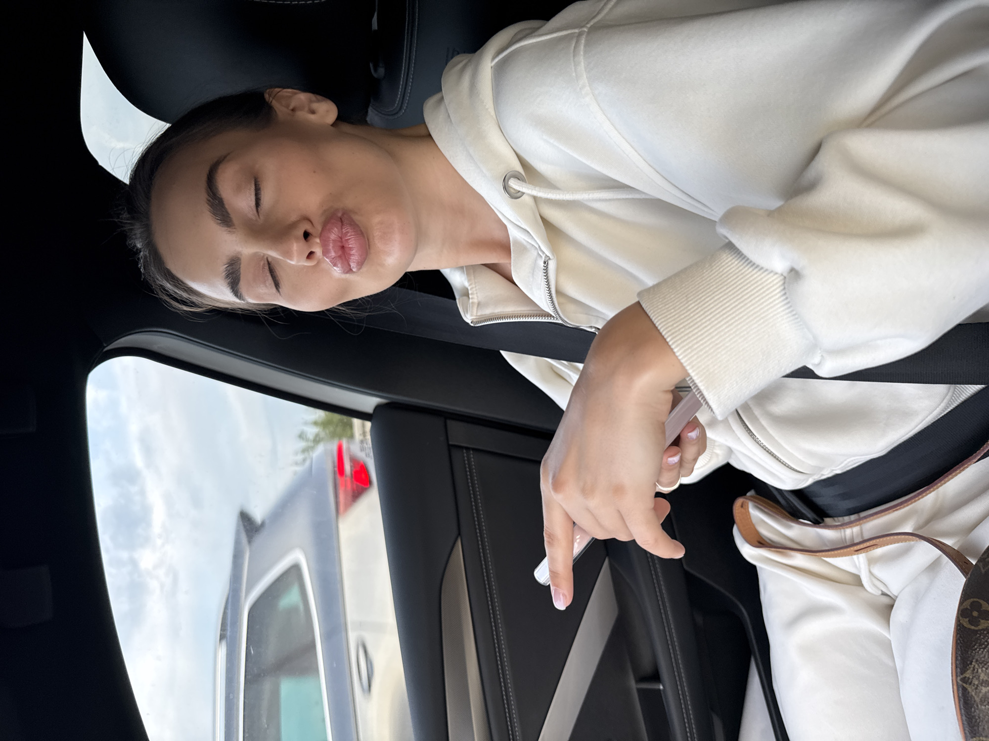 A woman wearing a white hoodie and a seatbelt is in a car. She is making a kissing face. Outside the car window is another vehicle. The city is unknown.