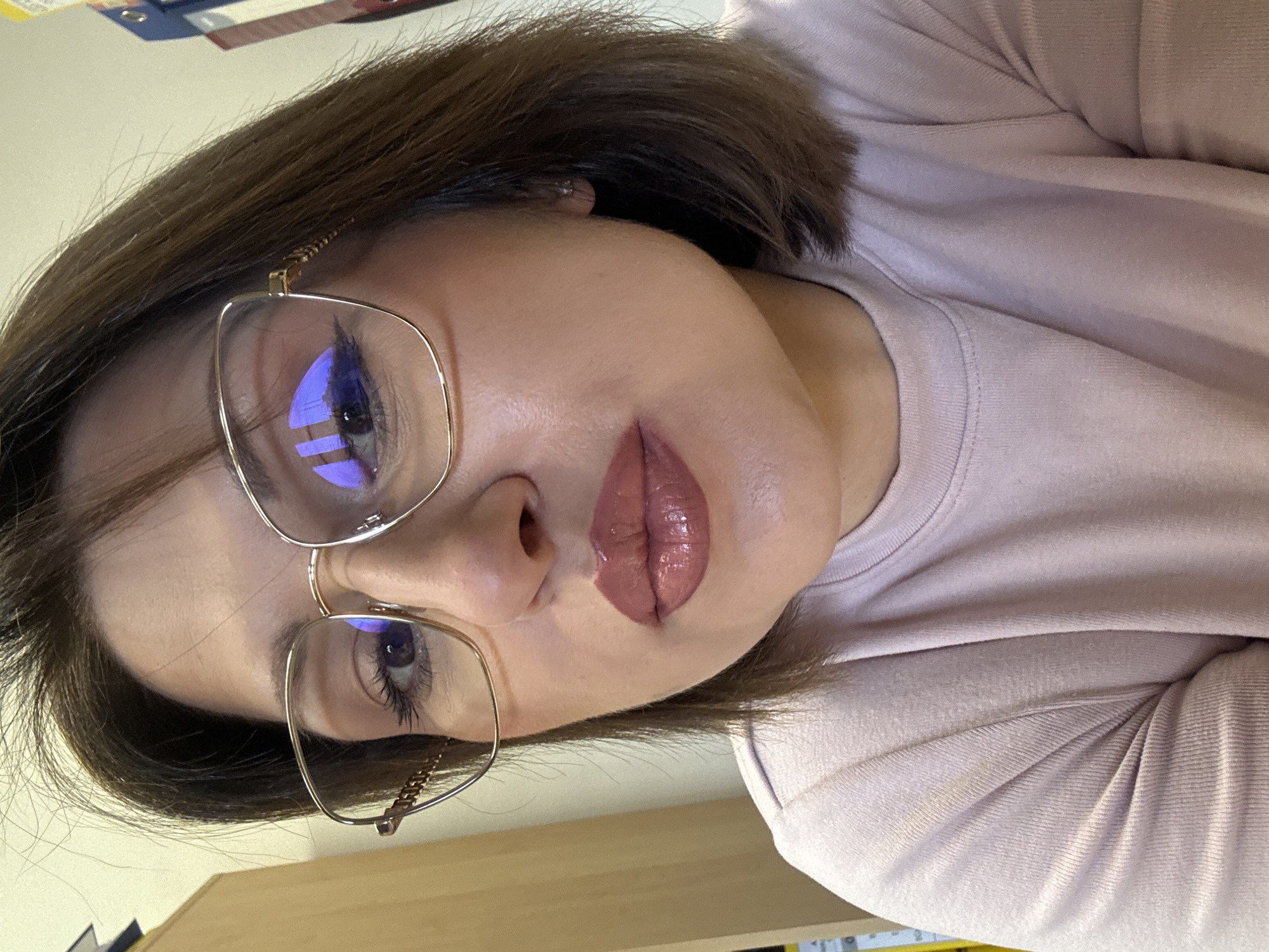 A woman is photographed in a close-up, slightly tilted angle. She wears glasses with gold frames. The reflection in her glasses reveals a cityscape, possibly in Sofia, Bulgaria. Her hair is short and dark. She is wearing a light pink sweater. Her lips are glossy and colored with a brownish-pink shade. The background is indistinct.