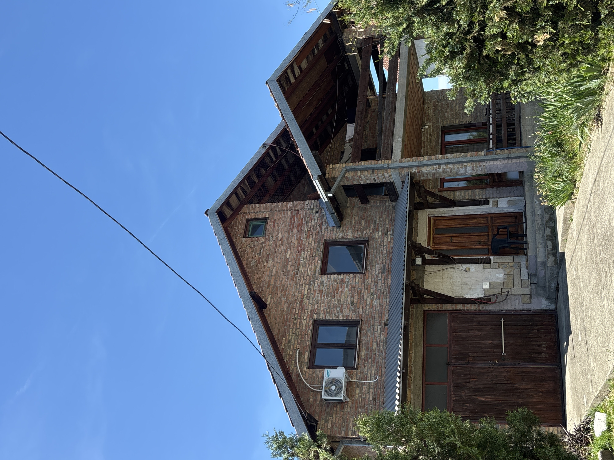 The image shows a two-story brick building in Grocka Urban Municipality, Serbia. The building has a partially exposed roof structure. There are several windows visible, and an air conditioning unit is mounted on the lower level. The building has a wooden door and a garage door. The sky is clear blue. A power line is also visible in the sky. Some vegetation is present in the foreground. A concrete structure is also visible.