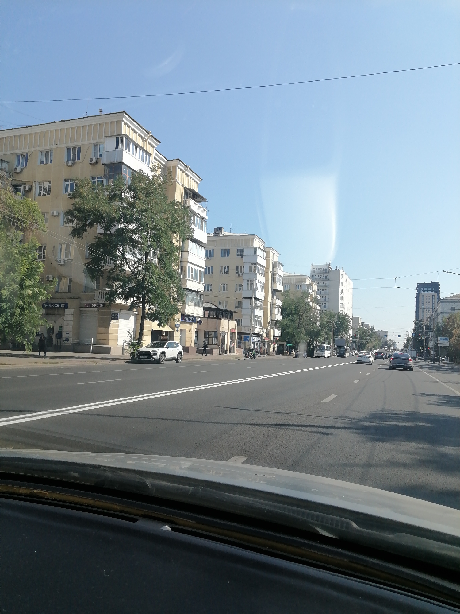 The image depicts a street scene in Rostov-on-Don, Russia, likely taken from inside a vehicle looking forward. The perspective is from the driver's seat, with the dashboard and windshield frame visible in the foreground. The street is a multi-lane asphalt road with clear white lane markings. Cars are driving in both directions. On the left side of the street, there are several multi-story buildings. The first building is light yellow with white trim and numerous windows, some with air conditioning units. Trees with green foliage are situated in front of this building and along the sidewalk. Further down the street on the left, there are more buildings, some with shops or businesses on the ground floor, indicated by signage. One sign visible says "Wildberries". On the right side of the street, there are also multi-story buildings, including a taller, modern building with a dark top. The sky is clear and blue, with some faint light streaks or atmospheric phenomena visible. The overall scene suggests a typical urban environment on a sunny day.
