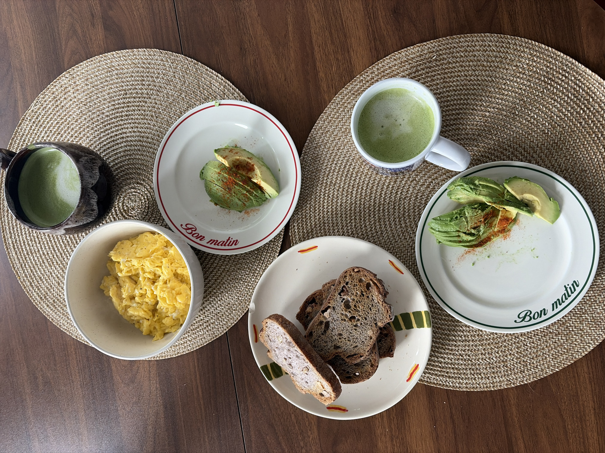 A high-angle shot captures a breakfast scene on a wooden table. Two circular woven placemats are positioned on the table. The breakfast consists of two plates with avocado slices sprinkled with red powder, a bowl of scrambled eggs, slices of dark bread, and two mugs filled with a green beverage, likely matcha. The plates feature the phrase "Bon matin," suggesting a French-Canadian origin, potentially Montreal, Canada.