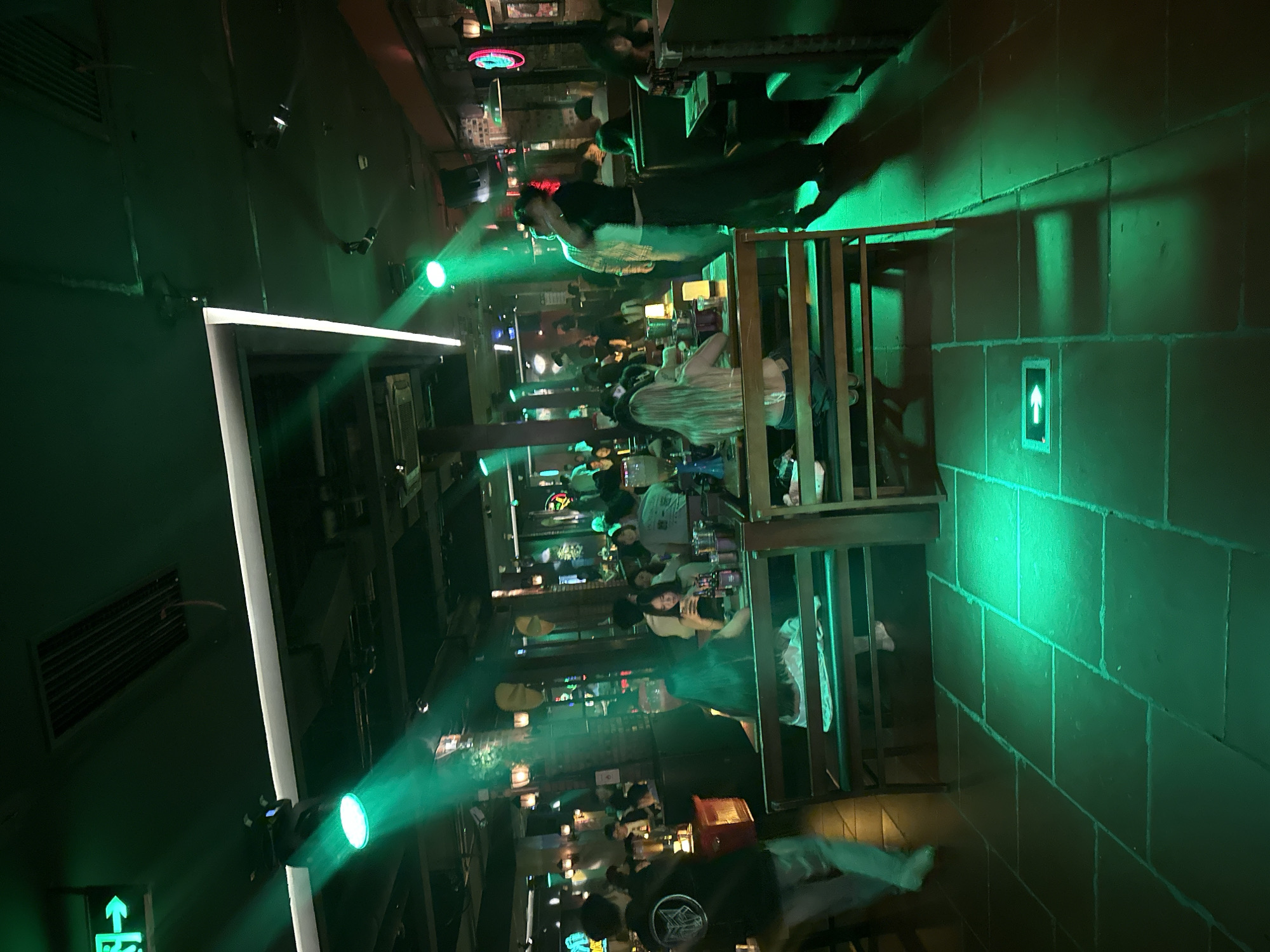The image depicts an indoor restaurant or bar environment illuminated with green lighting. Tables are occupied by numerous individuals, many of whom appear to be conversing or dining. A wooden railing is visible, possibly indicating a split-level layout. The setting appears to be a bustling establishment with a vibrant atmosphere. Location is presumed to be in Liangjiang, China, though specific landmarks or features confirming this are not evident in the image.