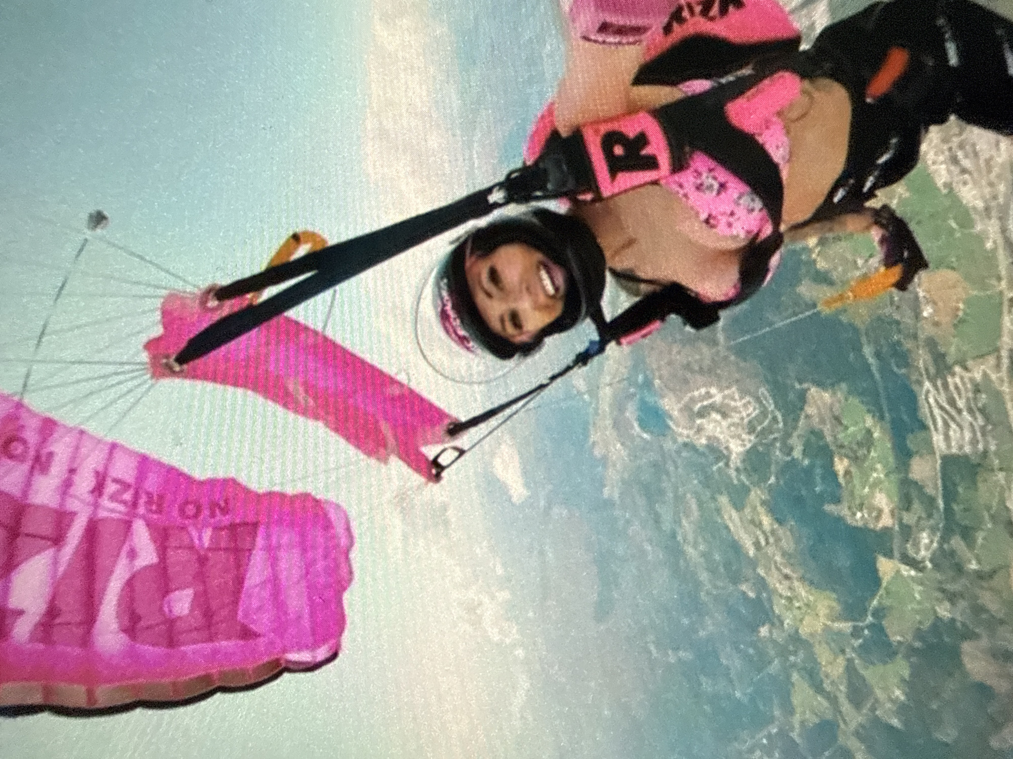 A woman skydives over an unidentified city. The woman has dark hair and is wearing a helmet. She wears a pink-and-black outfit. A pink parachute, emblazoned with the words "NO RIZK NO", is deployed above her. Below her, the terrain is visible, with water and landforms featuring patches of green.
