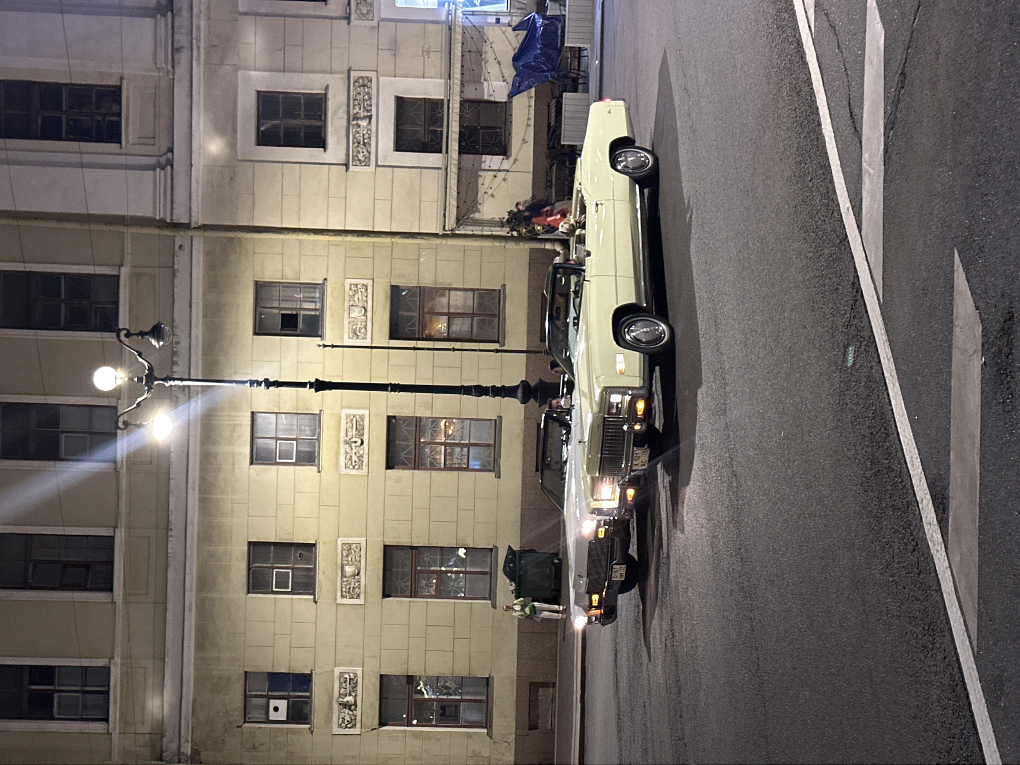 In an urban setting at night, two classic convertible cars, a white one and a pale yellow one, are parked on an asphalt road in front of a multi-story building. The building, illuminated by streetlights and interior lights, has a light-colored facade with several windows on each level. The white convertible is positioned slightly behind and to the left of the yellow convertible. The yellow convertible has its top down, and two people, a driver and a passenger, are visible inside. The road has visible white lane markings. A lamppost with two globes stands on the left side of the image, casting light. To the left of the cars, near the building's entrance, a dark green trash bin and a person in a light-colored outfit are present. The overall scene suggests a nighttime street view with vintage vehicles.