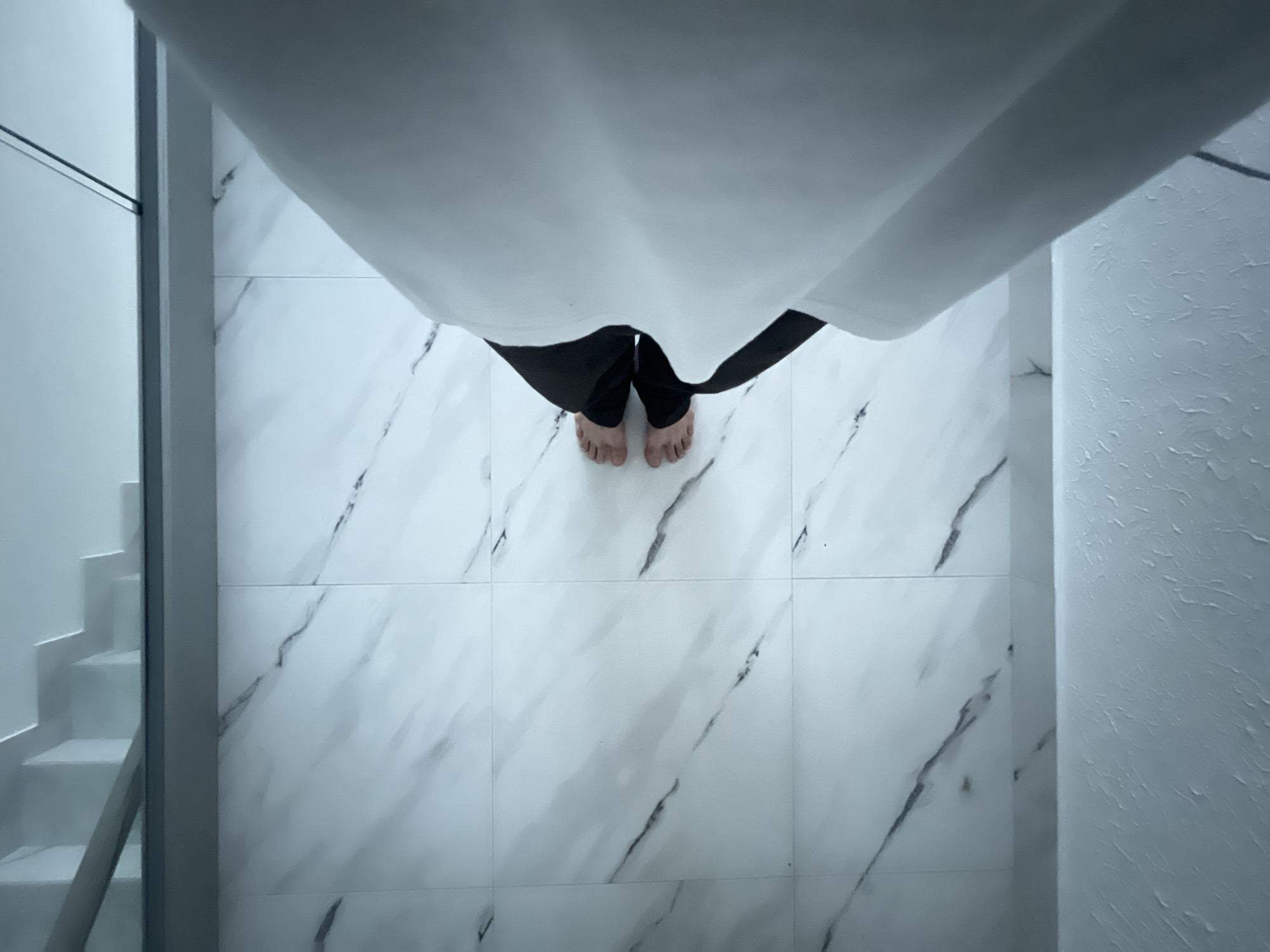 A top-down view captures a person standing barefoot on a white marble-tiled floor, partially obscured by a white cloth or curtain hanging from above. The person is wearing black pants. To the left, a white staircase is visible, and on the right, a textured white wall. The specific location in Sochi, Russia, cannot be determined from the given visual information.
