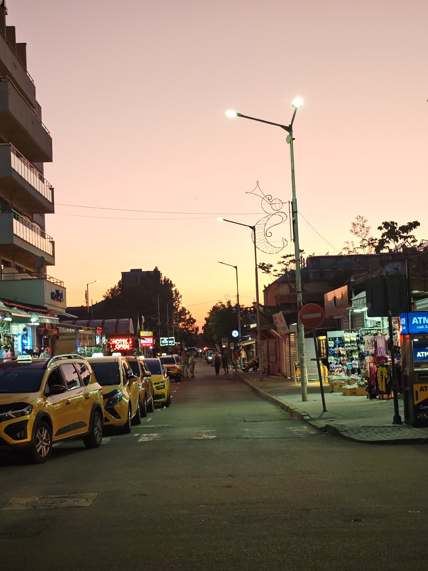 The image depicts a street in Nesebar, Bulgaria, at dusk. The sky transitions from orange to pink. Yellow taxis are parked on the left side of the road. Pedestrians walk in the distance. Stores line both sides of the street, illuminated by lights and neon signs such as "Hotel Open". A street light stands tall on the right, decorated with metal art. There is a “No Entry” traffic sign. An ATM is visible on the right. The street appears damp. A multi-story building is on the left side of the image.