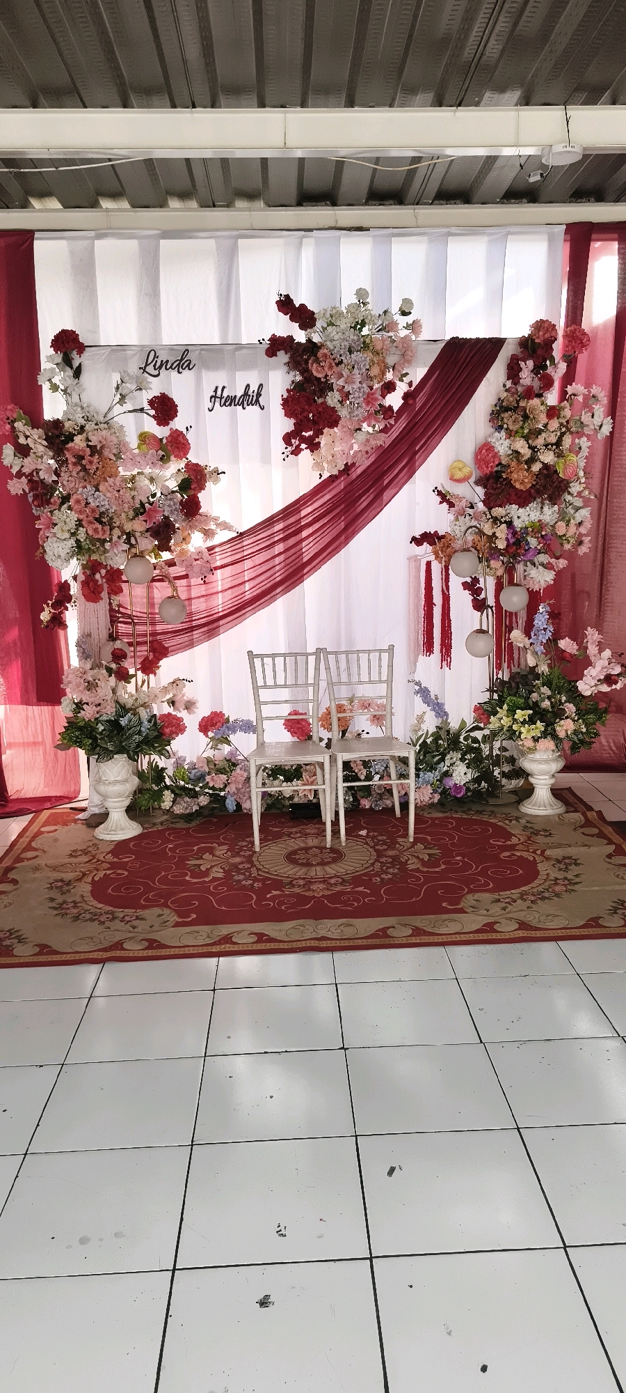 A wedding altar is set up indoors, likely at a venue in South Tangerang, Indonesia, given the style of decor common in the region. The altar features a white draped backdrop with maroon fabric swags. Two white chairs with decorative backs are placed on a red rug with an intricate floral pattern. The chairs face a floral arrangement that includes pink, white, and red flowers, with two decorative lamps on a stand to the left. The names "Linda" and "Hendrik" are visible in black cursive script on the backdrop. The floor is tiled in white.