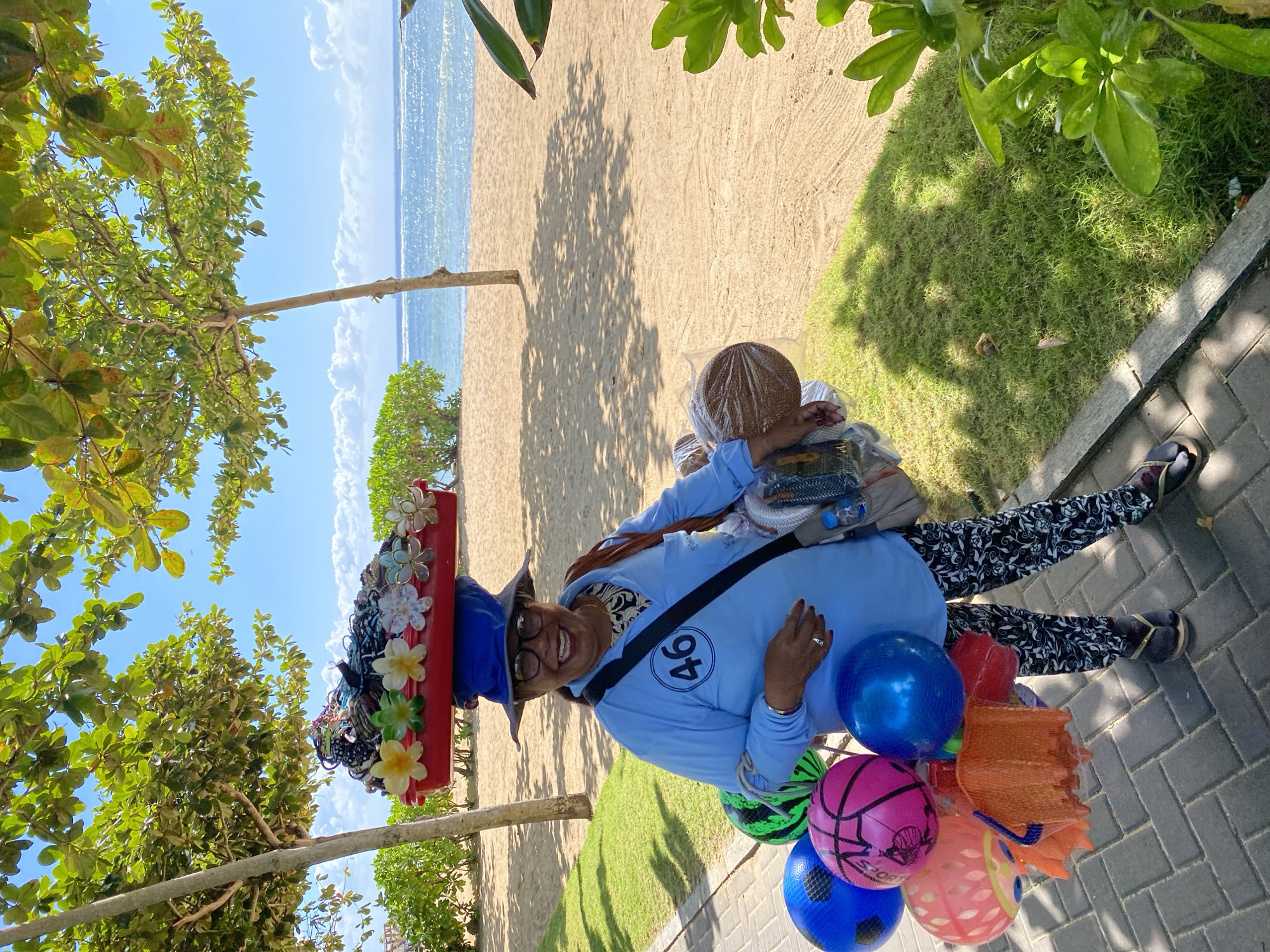 A woman in Kuta Selatan, Indonesia, stands smiling on a brick-paved area bordering a sandy beach. She wears a blue long-sleeved shirt with a "46" logo, patterned pants, and sandals. On her head, she has a large, decorated hat stacked with flowers and trinkets. She carries a collection of colorful balloons in one hand and a basket-like object covered in plastic wrap. The beach and ocean are visible in the background, framed by trees and greenery. The scene is bathed in sunlight.