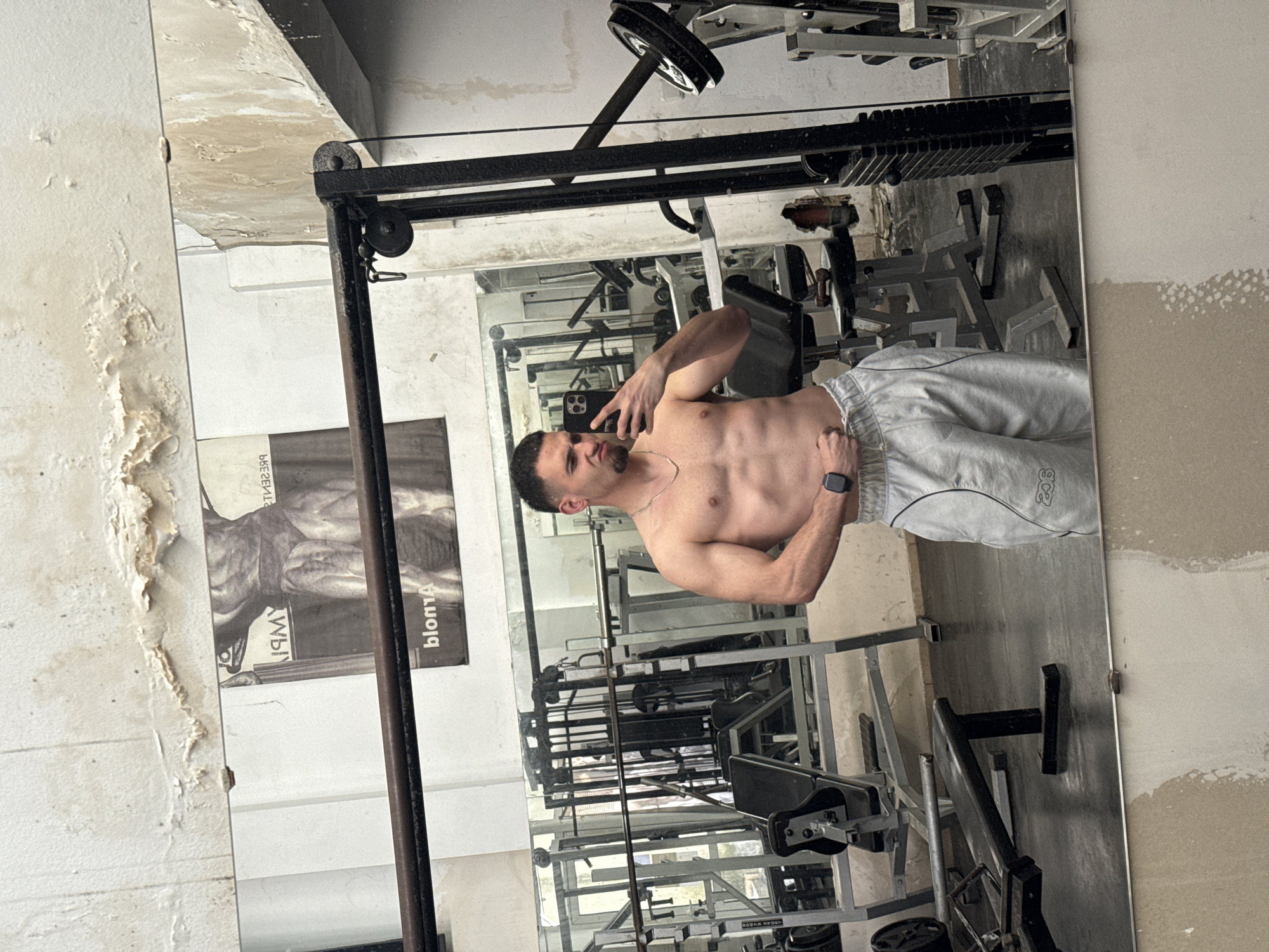 The image depicts a man taking a mirror selfie in a gym located in Shumen, Bulgaria. He stands shirtless, facing slightly right with a posed expression, looking towards his black smartphone which he holds up to capture the picture. He wears light grey sweatpants with the number "228" visible on his right thigh, a silver chain around his neck, and a dark smartwatch on his left wrist.
The gym environment appears aged or unmaintained. The walls, particularly to the left and top of the frame, exhibit significant peeling paint and damp stains, revealing the underlying plaster. Various gym equipment, including weight machines, dumbbells, and racks, are reflected in the large, full-body mirror behind the man. A black cable machine with a pulley system is prominently reflected in the foreground.
To the left of the mirror, a faded black and white poster depicts a muscular, possibly anatomical, figure. Partially visible reversed text on the poster includes "bloma" and "ЯМY". The scene captures a moment of self-documentation in a rustic fitness setting. The image is taken indoors, with no discernible weather details.