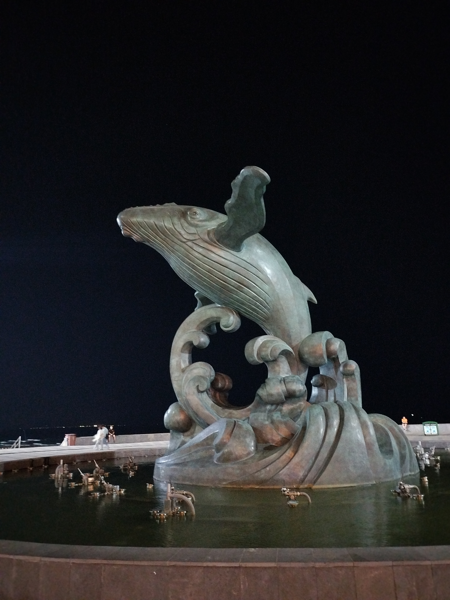 The image shows a whale statue at night in what appears to be a fountain. The statue is made of a grey-green material, possibly bronze or a similar metal, and depicts a whale emerging from waves. The fountain has multiple small water features that suggest a dynamic water display. In the background, there is a dimly lit walkway where people are visible, indicating a public space. Given the description, this statue is likely located in a coastal area in Ho Chi Minh City, Vietnam, as the city has coastal zones and a history of whale worship.