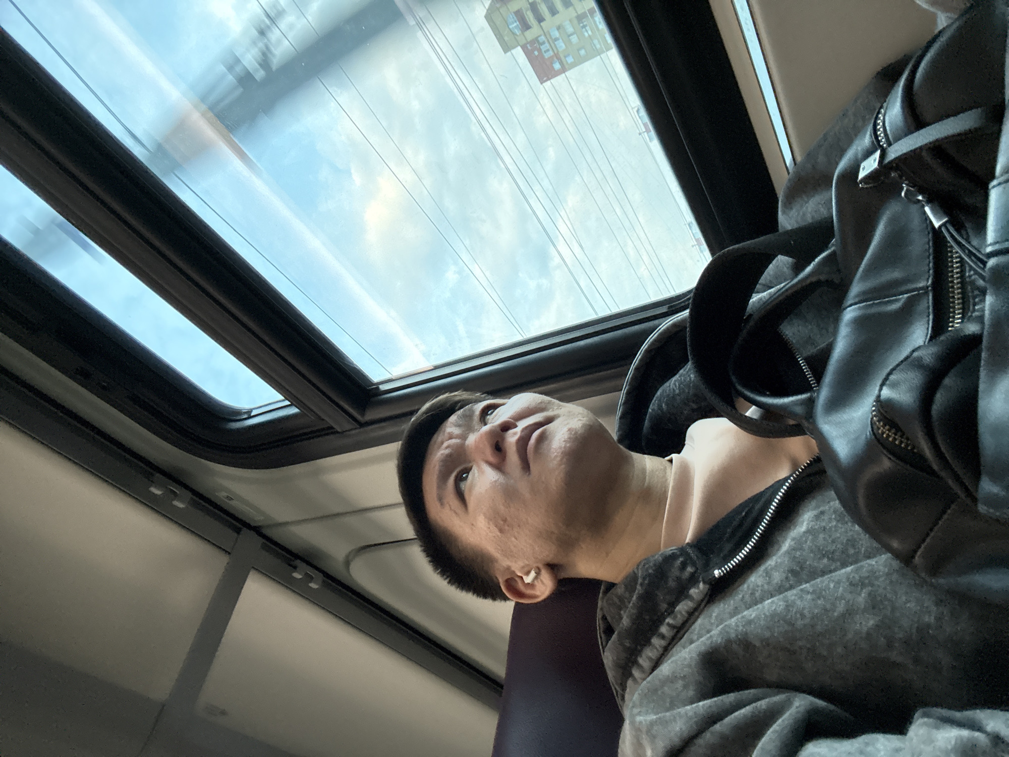 A low-angle view depicts an individual seated, gazing upwards. The person has short dark hair, light skin, and is wearing a white earbud in their left ear. They are clothed in a gray hoodie over a light-colored undershirt. A black leather bag rests to their right. Overhead, a window reveals a view of the sky, through which power lines are visible. A building with a red facade can be partially seen through the window. The location is in Unknown, Unknown.