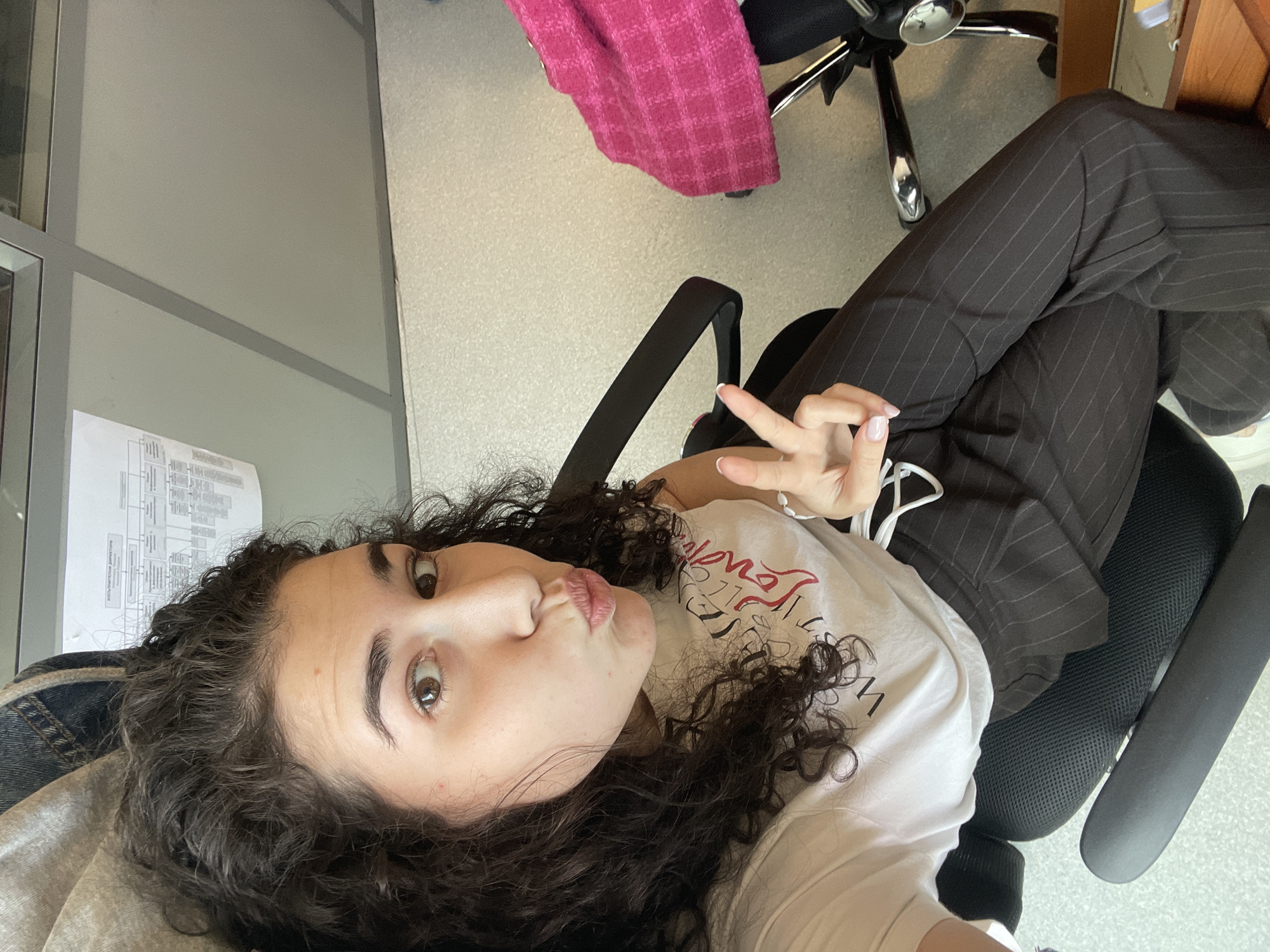 A selfie image, rotated 90 degrees clockwise, captures a young individual indoors, likely in an office or institutional setting. The person, with dark, voluminous curly hair and brown eyes, looks directly at the camera with a playful expression, making a "fish-lips" or puckered face. Their right hand is raised, showing a peace sign gesture, and a white wired earbud/headphone is visible near their hand.
The individual is wearing a white t-shirt with stylized red and black text; "WEST" is clearly visible in black, along with some red, partially obscured text and black scribbled designs. They are also wearing dark pinstriped trousers. A denim or textured jacket/shirt is draped over their left shoulder and arm. Their nails are manicured with a light polish and white tips.
The background reveals an office environment with light-colored flooring. To the left, a wall features large grey frames creating rectangular sections, resembling partitions or window panes. A white paper with diagrams and unreadable text is taped to this wall behind the person's head. To the right, a black office chair with a mesh back and chrome base is visible, along with a bright pink, textured fabric (possibly a jacket or blanket) draped over another chair or object in the distance. A wooden desk or cabinet corner is also partially seen in the upper right. The lighting suggests it is daytime. The overall mood is casual and relaxed.