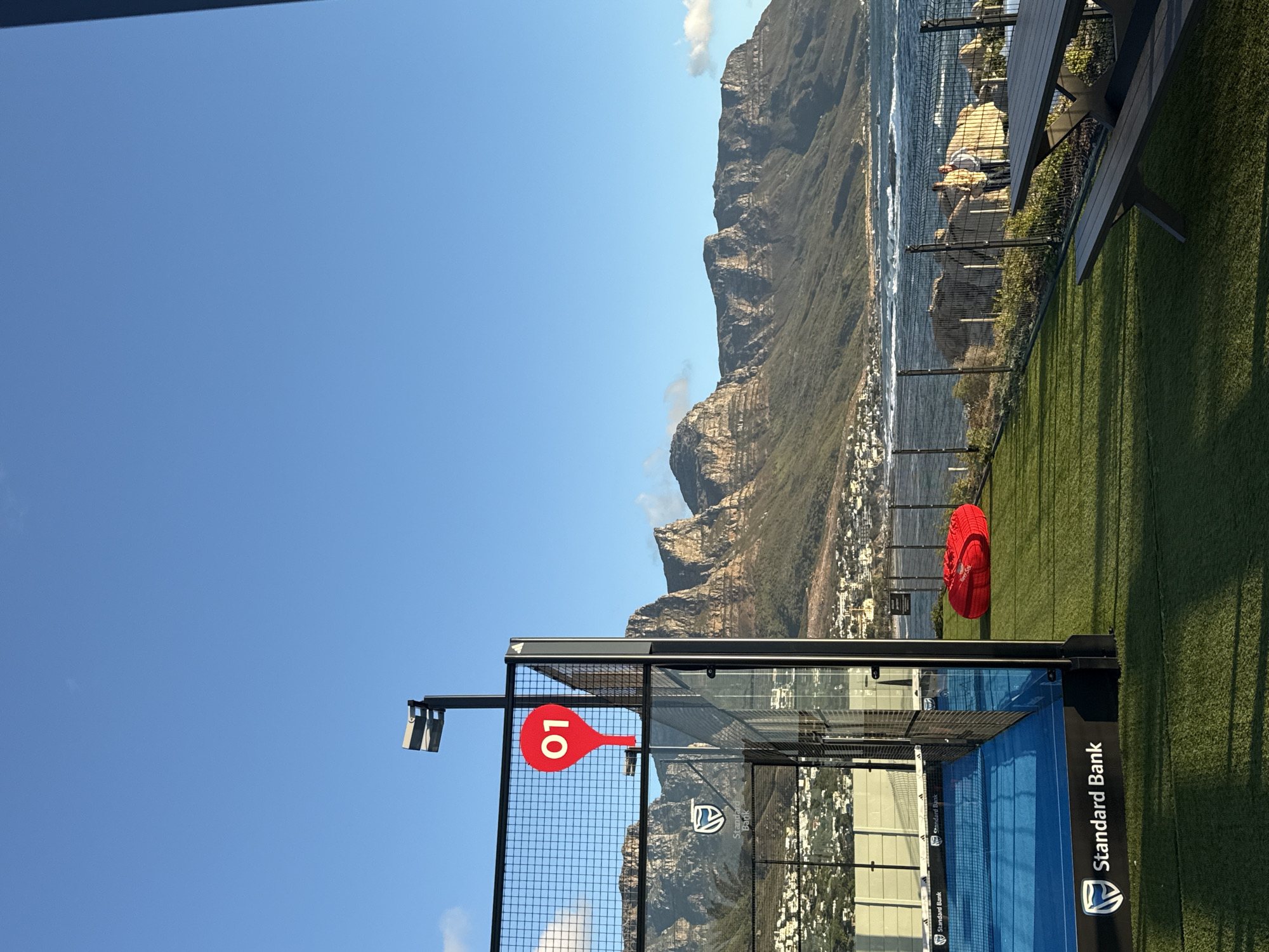 A clear day in Cape Town, South Africa, features a padel tennis court overlooking the Atlantic Ocean. The court, branded with "Standard Bank," has blue flooring and black fencing with a red paddle-shaped sign with the number "10." In the background, Table Mountain dominates the landscape. Above the court, a patch of artificial green grass is occupied by a red circular object, perhaps a cushion or beanbag, and a wooden picnic table with benches. Across the fence, two people are sitting by the ocean. The sky is a cloudless blue.