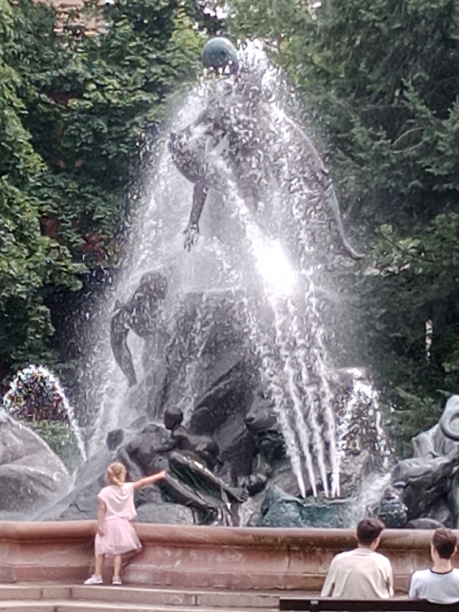 The image shows a large, ornate fountain with a girl in a pink dress standing on a ledge, pointing towards the fountain. The fountain features multiple sculpted figures and water cascading down. In the foreground, two young men are sitting on a bench, facing away from the camera. The fountain is surrounded by lush green trees. The scene appears to be a public park or square. There is no indication of the location being in Bydgoszcz, Poland.