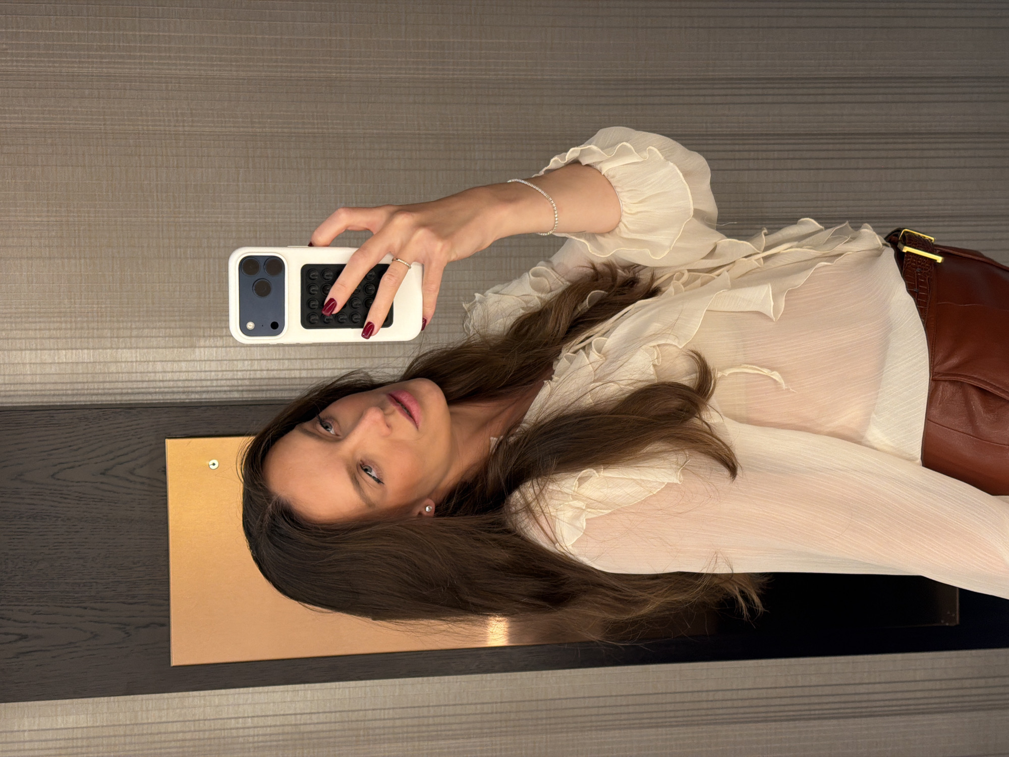 A woman with long brown hair is taking a selfie with her smartphone in what appears to be an interior setting. She is wearing an off-white blouse with ruffles and a brown leather belt. Her nails are painted dark red, and she has a silver bracelet on her left wrist. A corner of what seems to be a dresser or table with a gold panel is visible. The background consists of horizontally striped wall panels. The location in Budva, Montenegro is not determinable from the visual information.