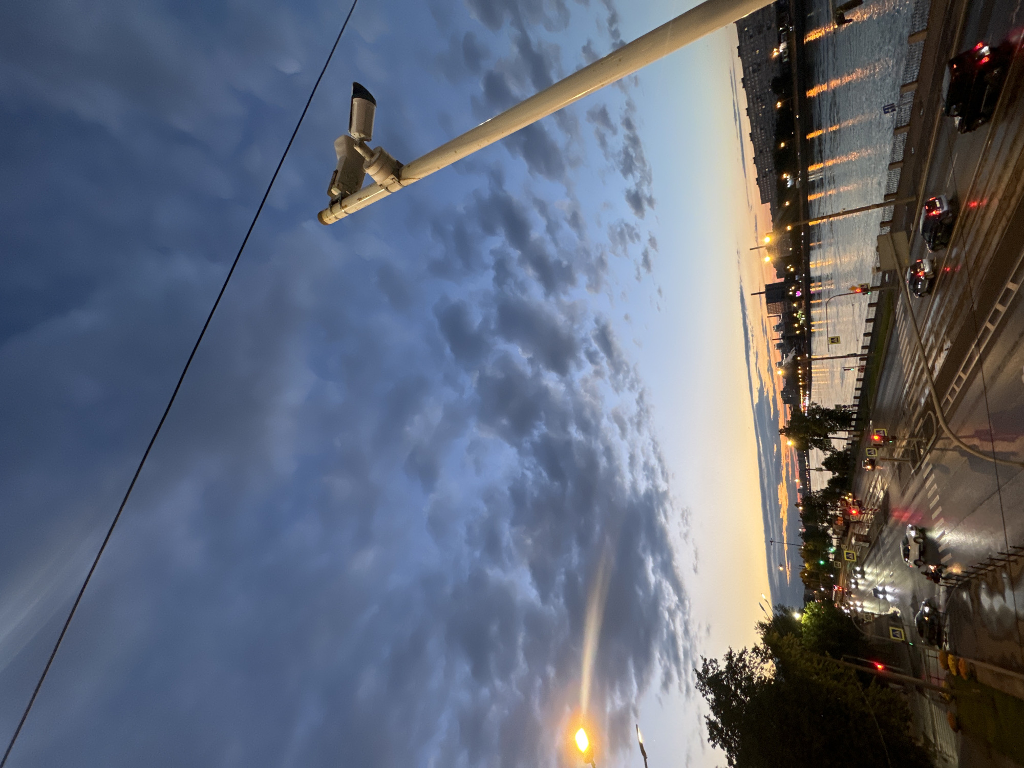 The image captures a twilight scene over a wide river in Kudrovo, Russia. The sky is a dramatic mix of deep blues and grays, with scattered clouds, illuminated by the remnants of sunset. A prominent utility pole with a security camera and a light fixture stands on the right, angled towards the sky. Below, a wet, multi-lane road curves along the riverbank, reflecting the lights of the city and traffic signals. Several cars are visible on the road, moving in various directions. Pedestrian crosswalks are clearly marked. Trees line the left side of the road, and buildings form a cityscape across the river. The overall atmosphere is one of urban tranquility at dusk.