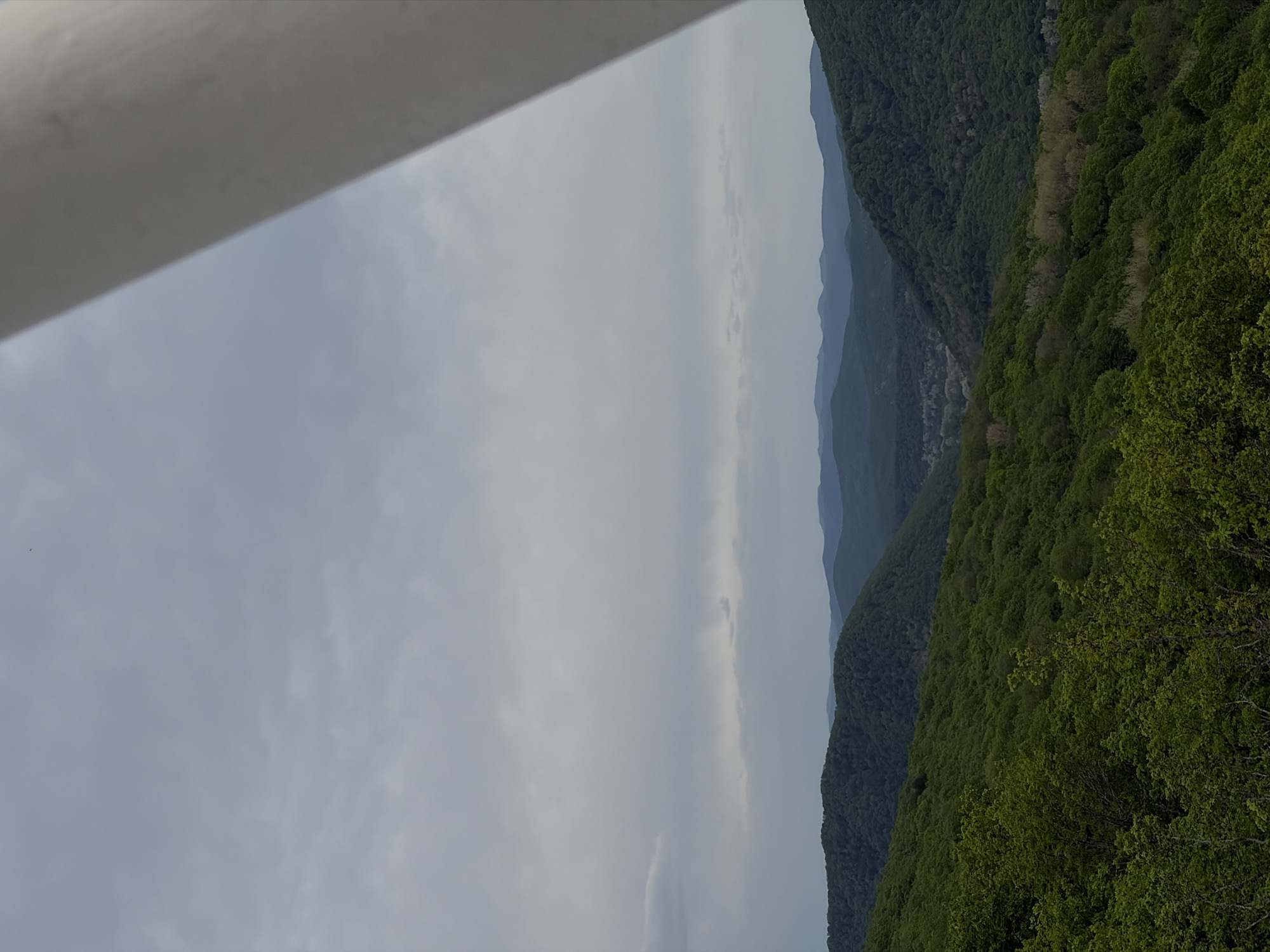 A landscape photo taken in Goryachiy Klyuch, Russia, shows a view from an elevated position looking across a valley filled with lush, green forested hills. In the distance, other ridges of mountains are visible. The sky is overcast, with a pale grey tone. A portion of a building or structure is visible in the upper left corner of the frame.