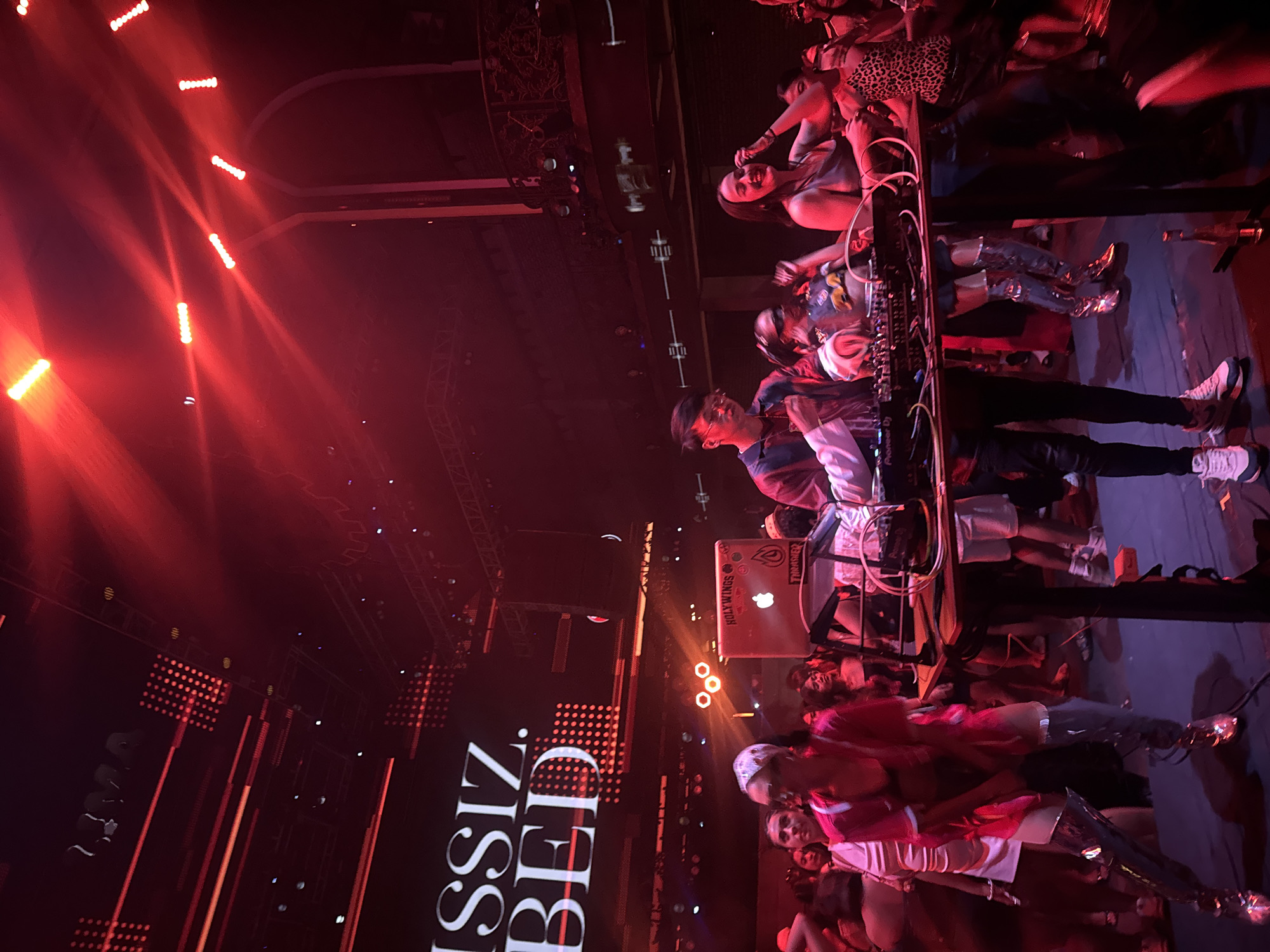 The image captures a vibrant nighttime scene at a club or event venue, likely in North Kuta, Indonesia. A DJ is actively performing, with a DJ setup including a laptop, mixer, and various electronic equipment prominently displayed. The DJ is surrounded by a lively crowd of people, many of whom appear to be dancing and enjoying the music. The atmosphere is illuminated by dramatic red stage lighting, casting a warm and energetic glow across the scene.
The crowd consists of individuals dressed in casual and stylish attire, with some notable fashion choices such as metallic boots. The DJ is wearing a face covering and a colorful outfit, adding to the festive mood. Some individuals in the foreground are looking towards the camera, while others are engaged in the activities around them. The background reveals more of the venue's infrastructure, including stage lighting rigs and decorative elements.
The visible text on the laptop reads "HOLY WINGS" and "THRASHER." A large digital display in the background shows the stylized text "ASSISZ BED" in white against a dark background. The overall impression is one of a high-energy, entertainment-focused event, possibly a music performance or a club night, filled with excitement and a sense of community. The time of day is clearly night, and the indoor setting suggests a controlled environment for the event.