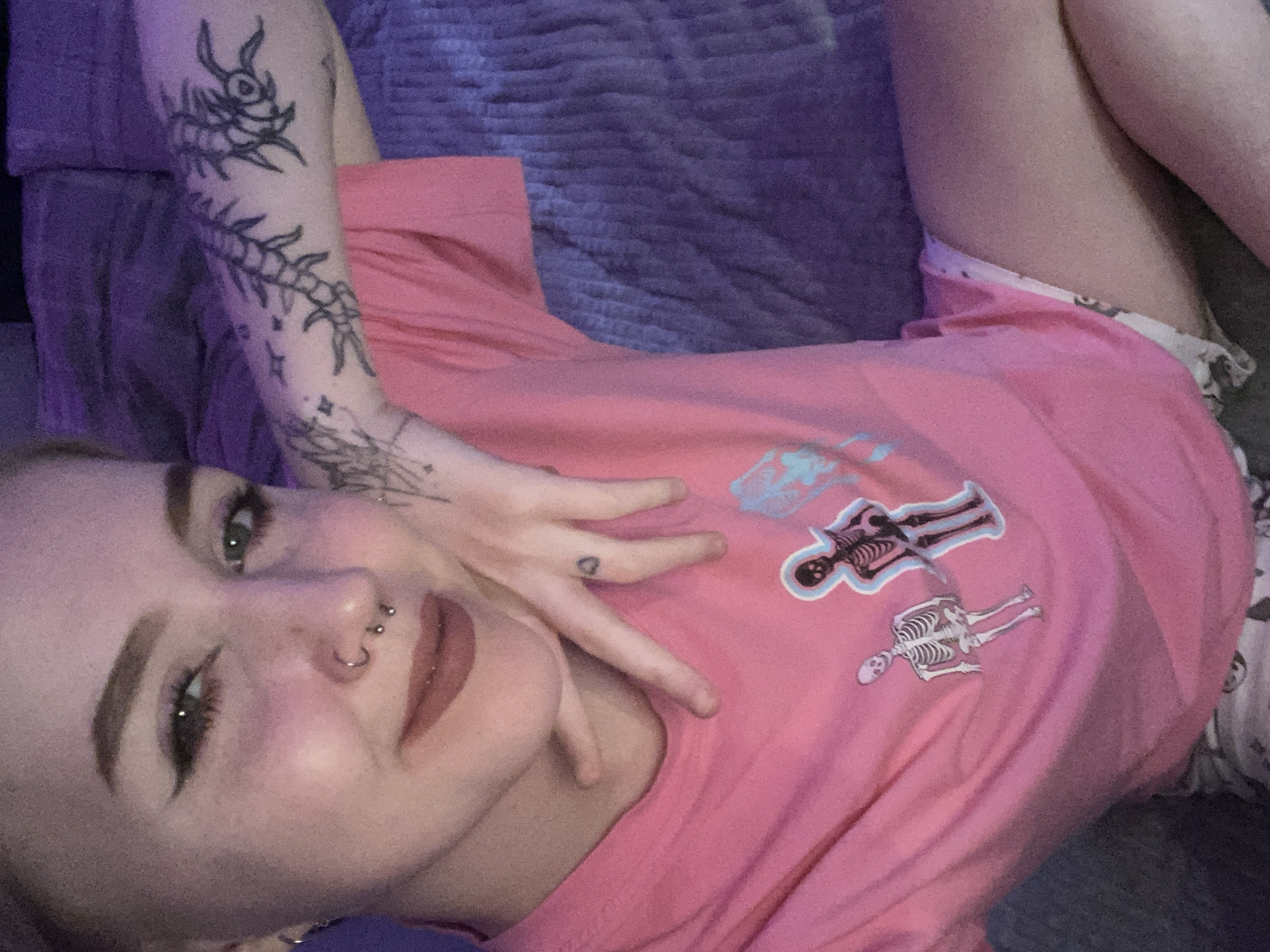 A woman lies on a cushioned surface, likely a couch or bed. She wears a pink t-shirt with skeleton motifs and patterned bottoms. She has arm tattoos and facial piercings. The background includes a textured, possibly quilted, surface. There is no evidence to suggest the location is Izhevsk, Russia.