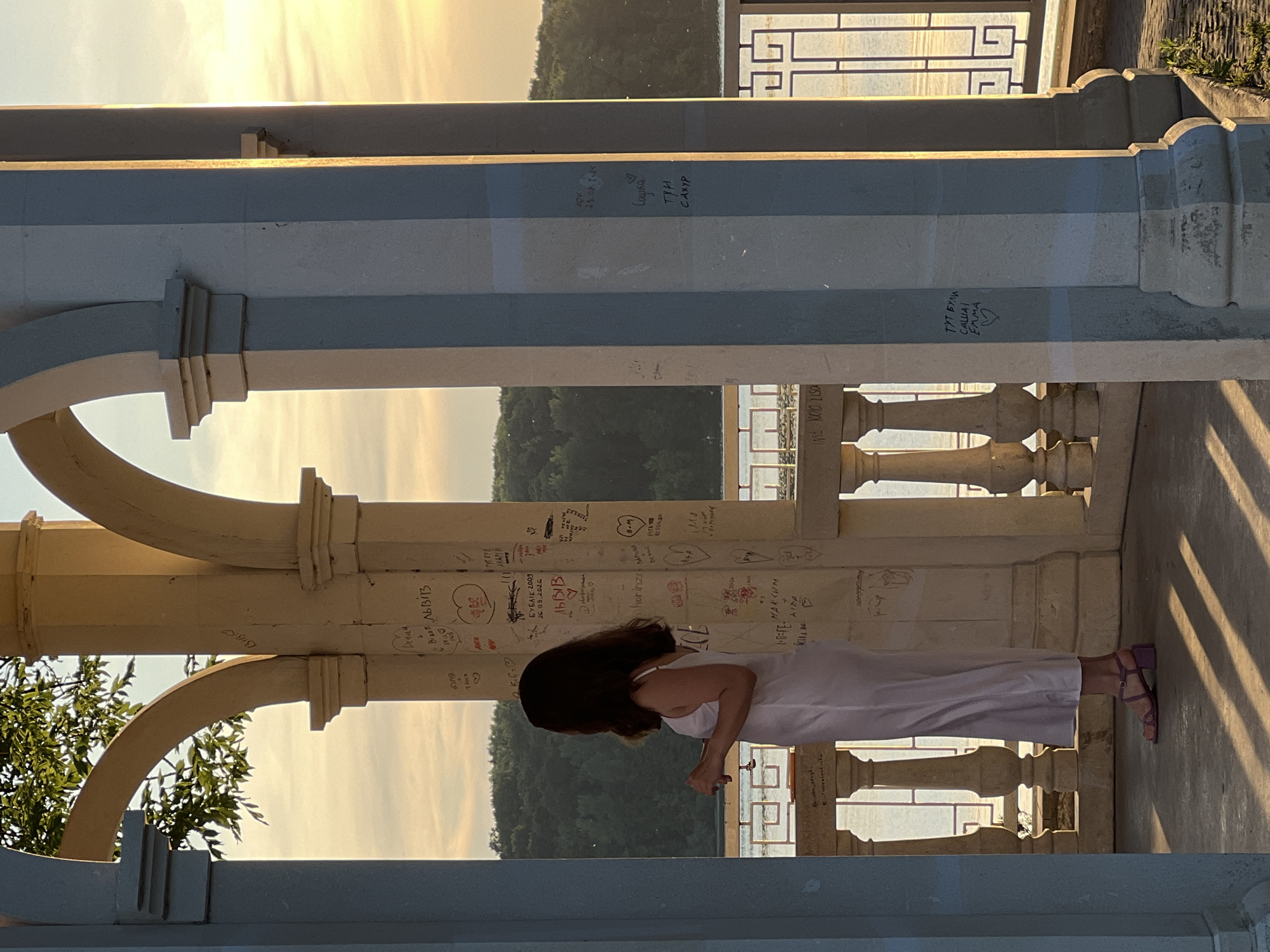 The image depicts a woman standing inside a stone gazebo with arches, facing away from the camera and towards a scenic view. The gazebo is adorned with graffiti. The woman is wearing a white slip dress and purple strappy heels. In the background, a body of water and a tree-covered hillside are visible under a warm, golden sunset sky. This is likely the Ternopil "Love Island" (Ostriv Kohannya) gazebo, a popular spot located on the embankment of the Ternopil Pond in Ternopil, Ukraine.