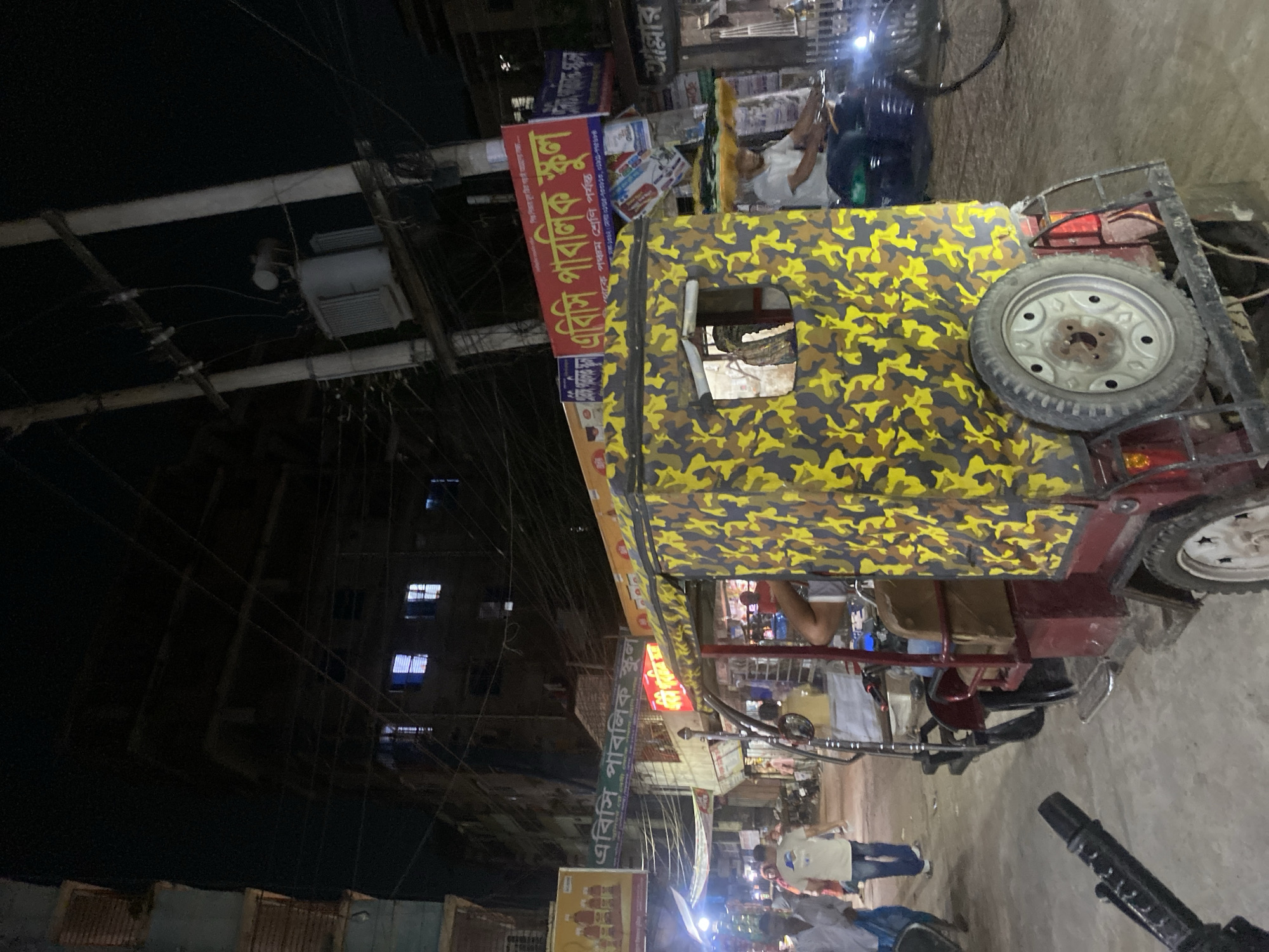 The image captures an urban scene at night, possibly in Dhaka, Bangladesh, featuring a distinctive three-wheeled motorized rickshaw prominently displayed. The rickshaw has a camouflage pattern and is laden with various items, including a spare tire mounted on the back.
A sign partially visible above the rickshaw reads "এবিসি পাবলিক স্কুল", indicating a local school, and includes phone numbers, suggesting a commercial aspect. Below this sign, another sign duplicates the information.
The background reveals a street scene with pedestrians, lights, and other vehicles, which contributes to the lively urban atmosphere. The image's tilt and perspective suggest it was taken spontaneously, capturing a slice of everyday life in a bustling South Asian city. Electrical wires are prominently visible in the upper left, adding to the sense of dense urban infrastructure.