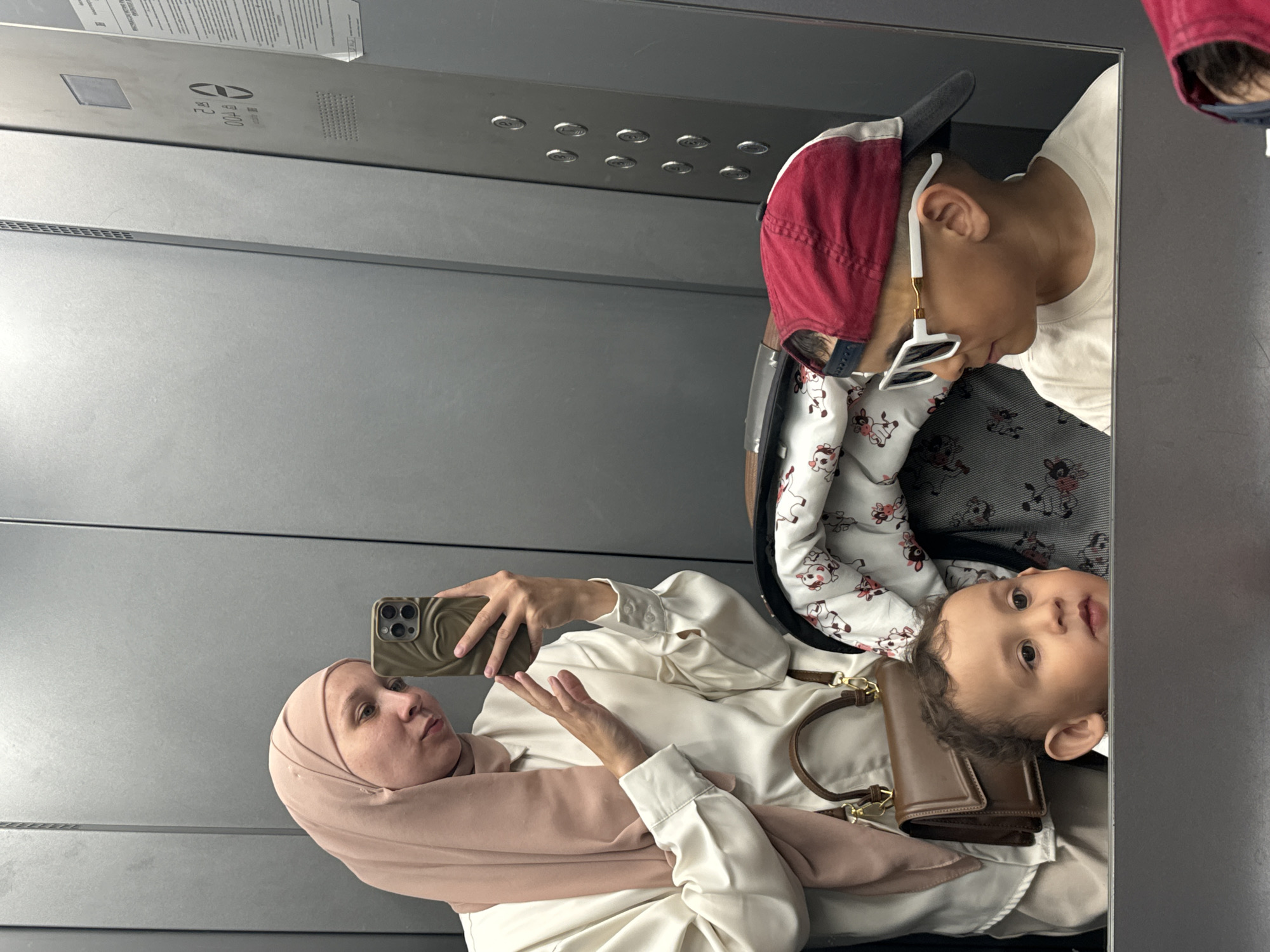 A woman wearing a peach-colored hijab and white blouse is taking a picture with her iPhone in what appears to be an elevator. Two children are also visible in the image. One child, with curly hair, is in a stroller or sitting. A second child is wearing a white t-shirt, a cow print jacket, and a red and gray baseball cap, and white sunglasses. The elevator interior is gray metal. The location in the city of Chaikovsky, Russia, is unknown.